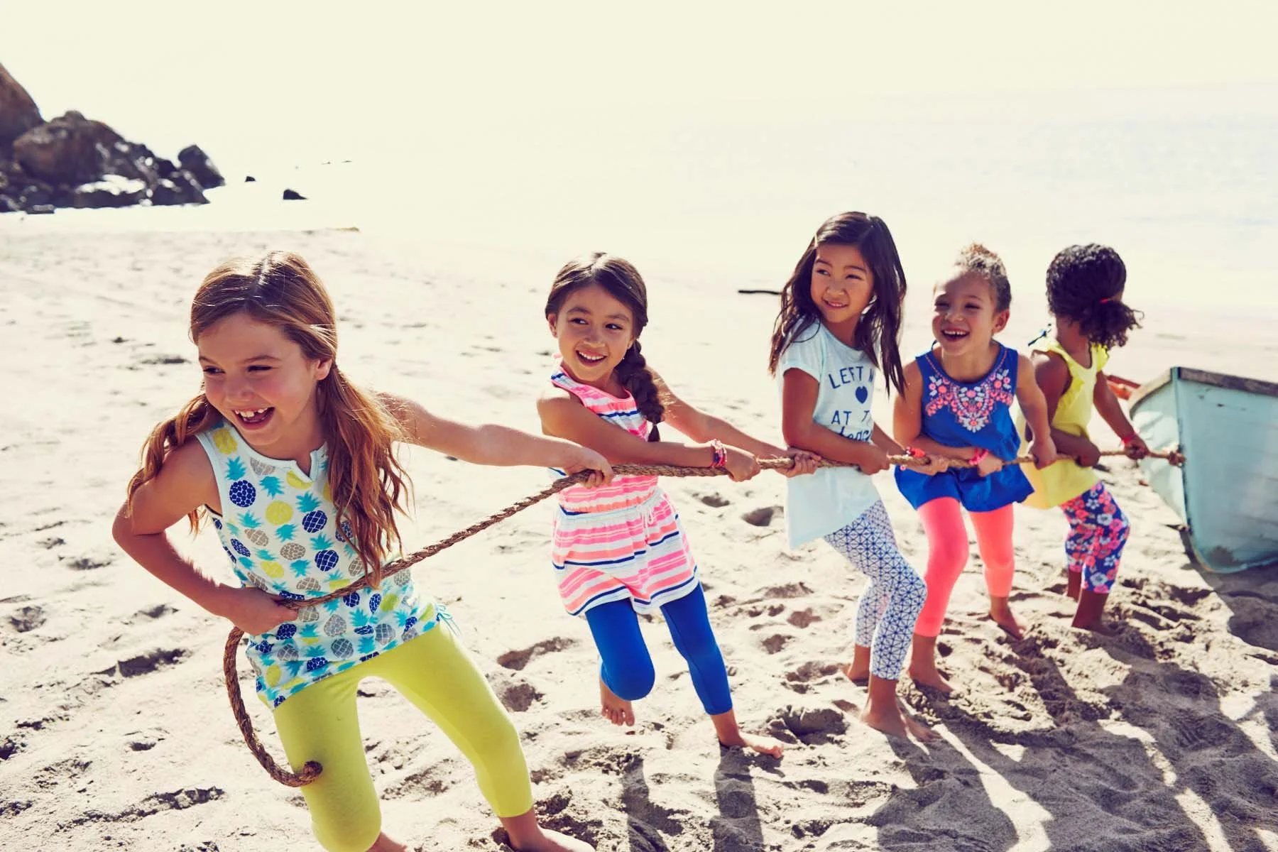 Five young girls with different hair colors and styles playing tug of war with a rope on a sunny beach, smiling and having fun.