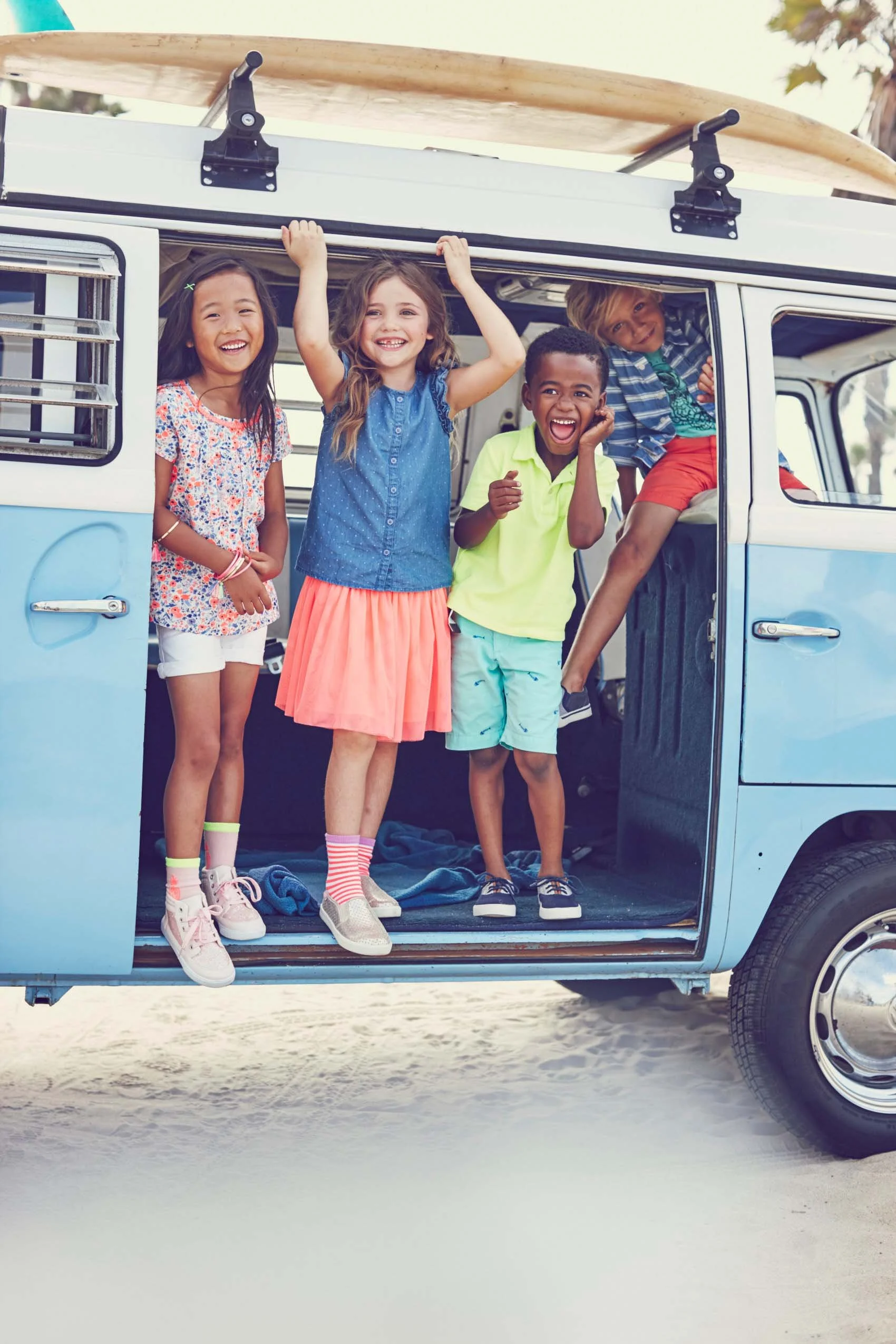 Four children smiling and laughing while standing inside a blue camper van with a surfboard on top, parked on a sandy beach.