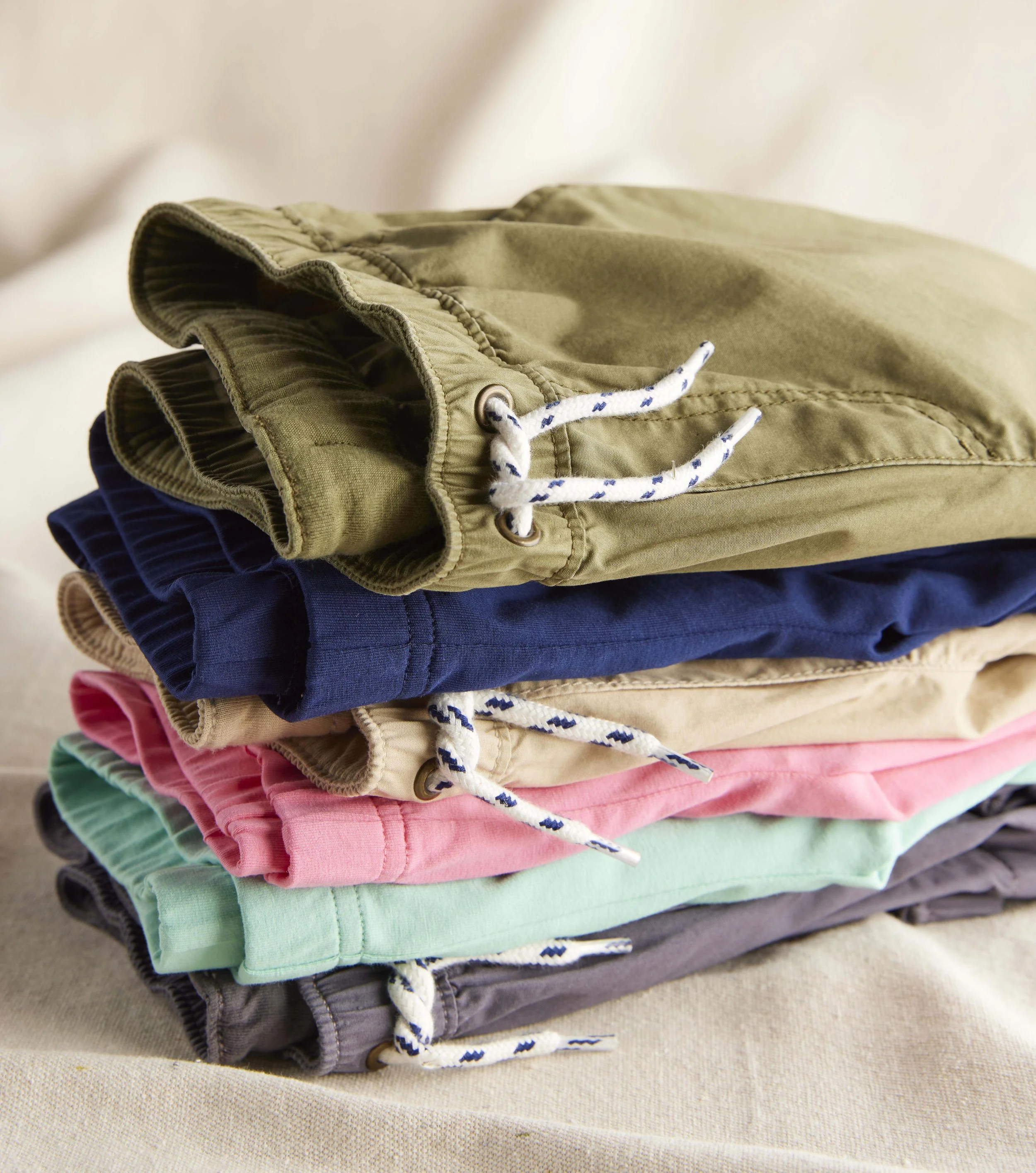 A stack of folded pants in different colors including green, pink, light green, gray, navy blue, beige, and black, placed on a beige fabric surface.