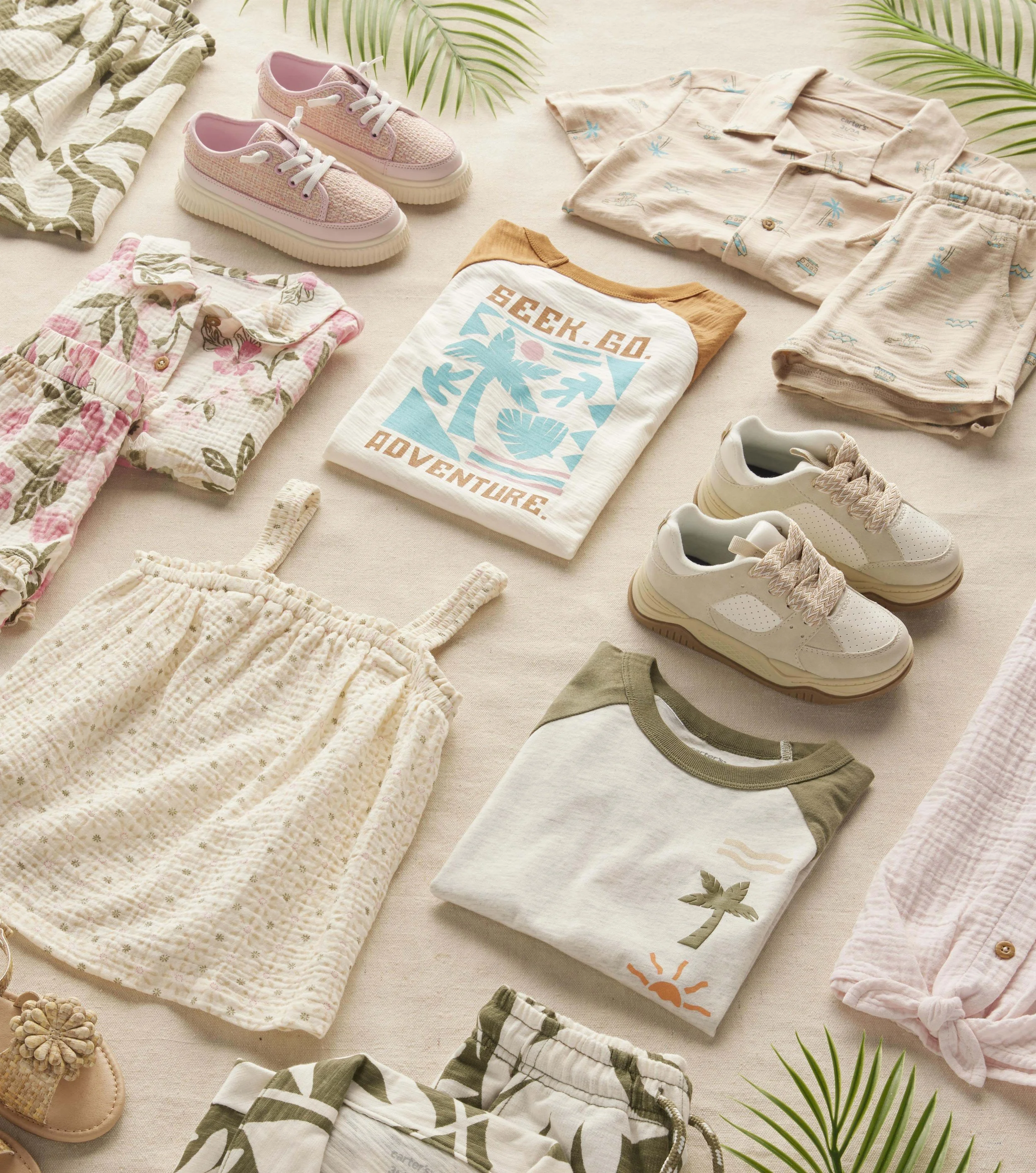 Flat lay of children's clothing and accessories on a light-colored surface, including floral pajamas, a graphic T-shirt with 'Beach. Go. Adventure.', patterned shorts, sneakers, and sand-colored shoes, with green plant leaves in the background.