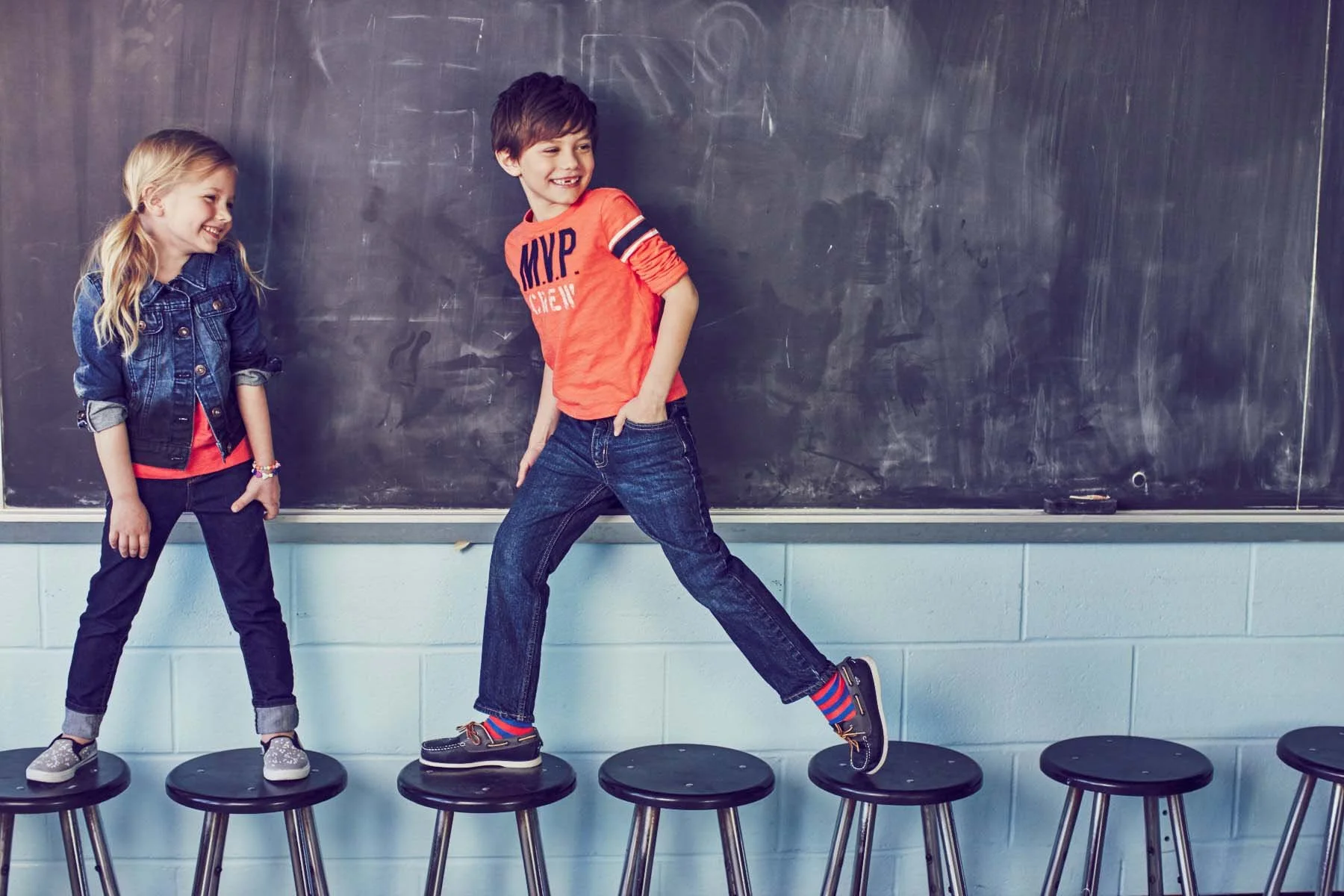 Three children standing on black round stools in front of a large chalkboard in a classroom. The girl on the left has blonde hair, wearing a denim jacket, black pants, and sneakers. The boy in the middle has brown hair, wearing an orange long sleeve 