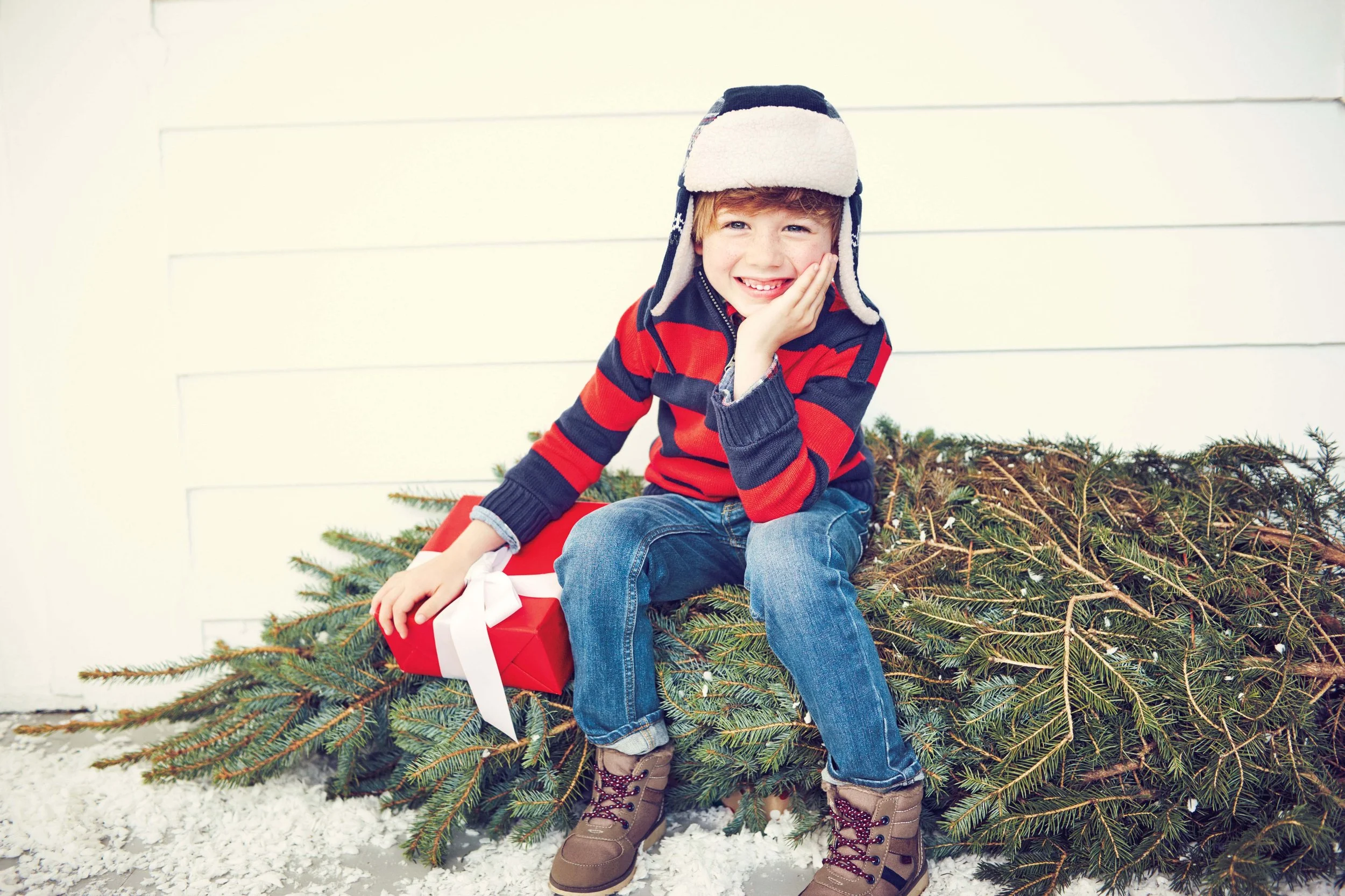 A young boy sitting on a Christmas tree decorated with ornaments, wearing a winter hat, red and navy striped sweater, blue jeans, and brown boots, smiling and touching his face with one hand, with a wrapped gift with a white ribbon nearby.