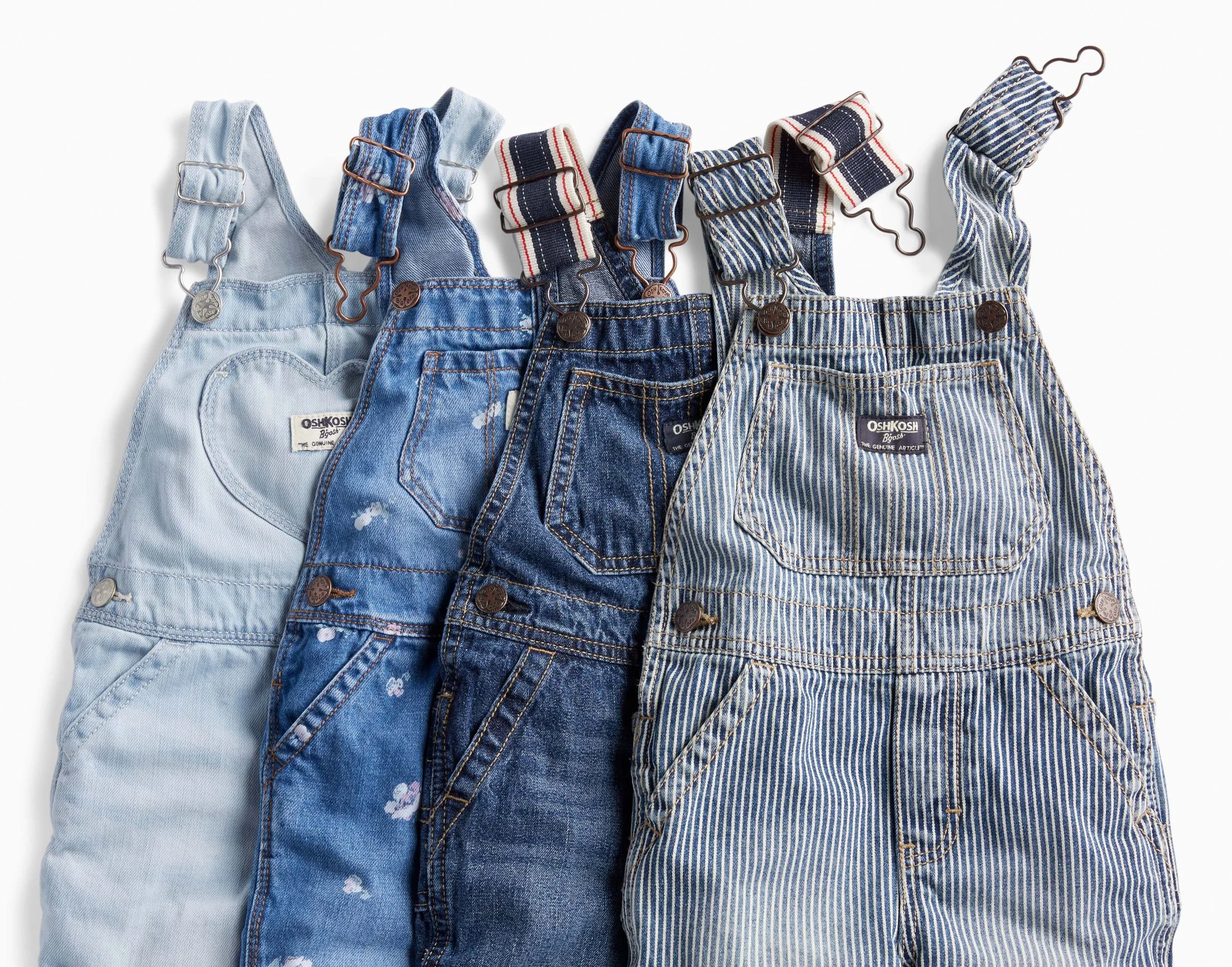 A row of seven denim overalls hanging on hooks against a white background, with different shades and styles of blue denim.