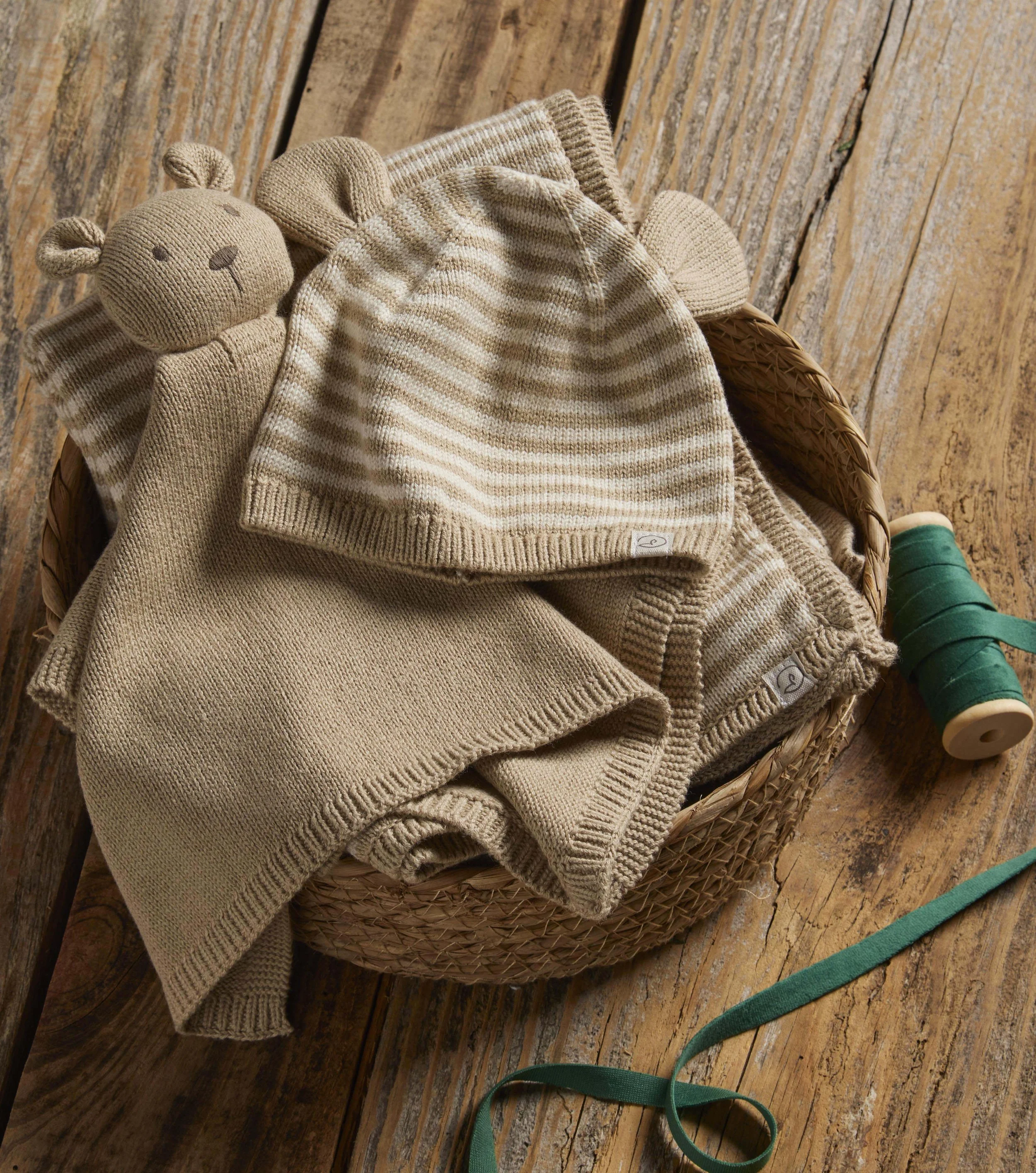 A knitted beige teddy bear with a round head, small ears, and a simple face, resting on a pile of beige and striped knitted baby clothing in a woven basket. The basket is on a rustic wooden surface, with some green ribbon or fabric nearby.