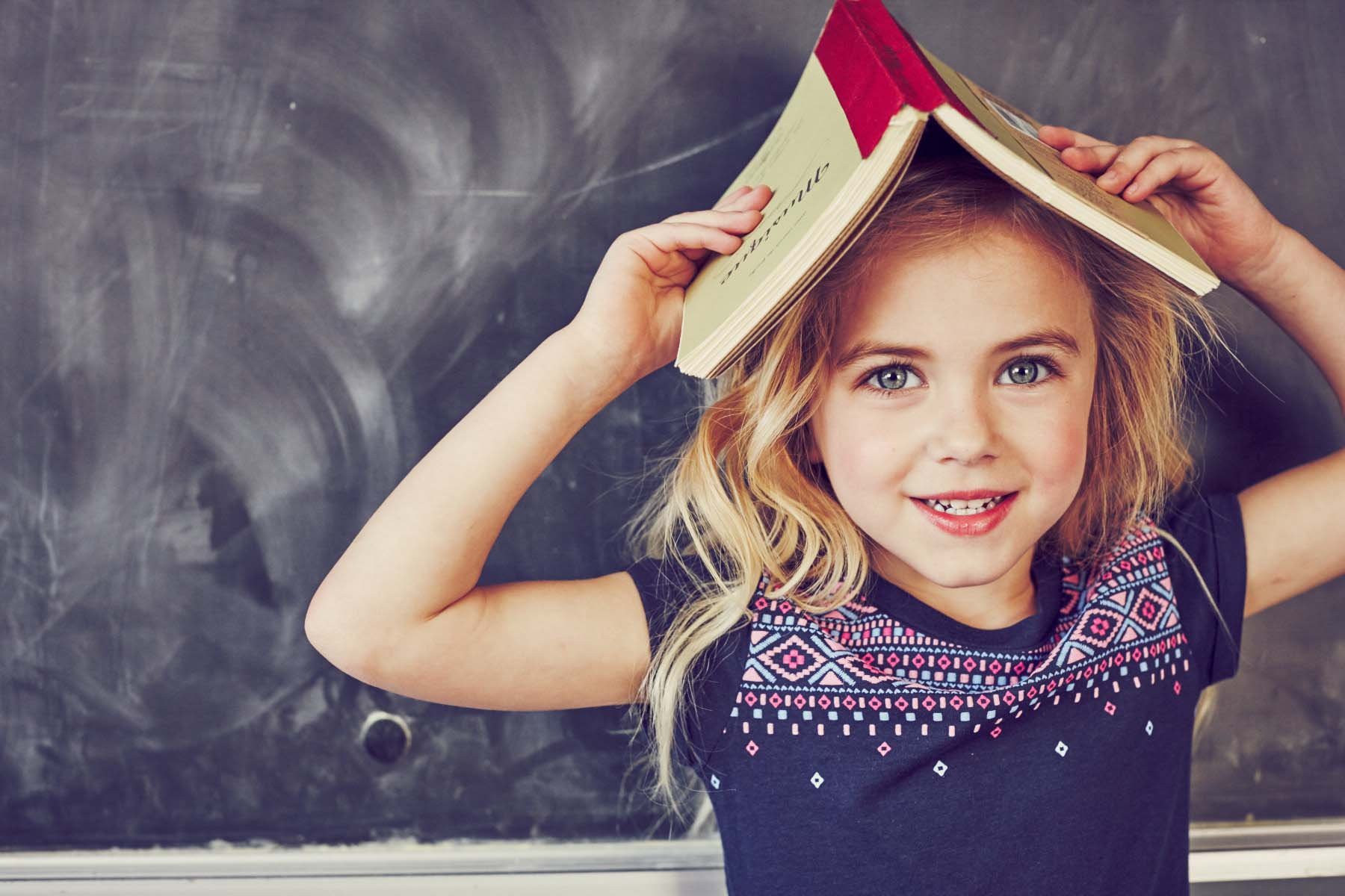 A young girl with blonde hair and blue eyes smiling, holding a book above her head like a hat, standing in front of a chalkboard.