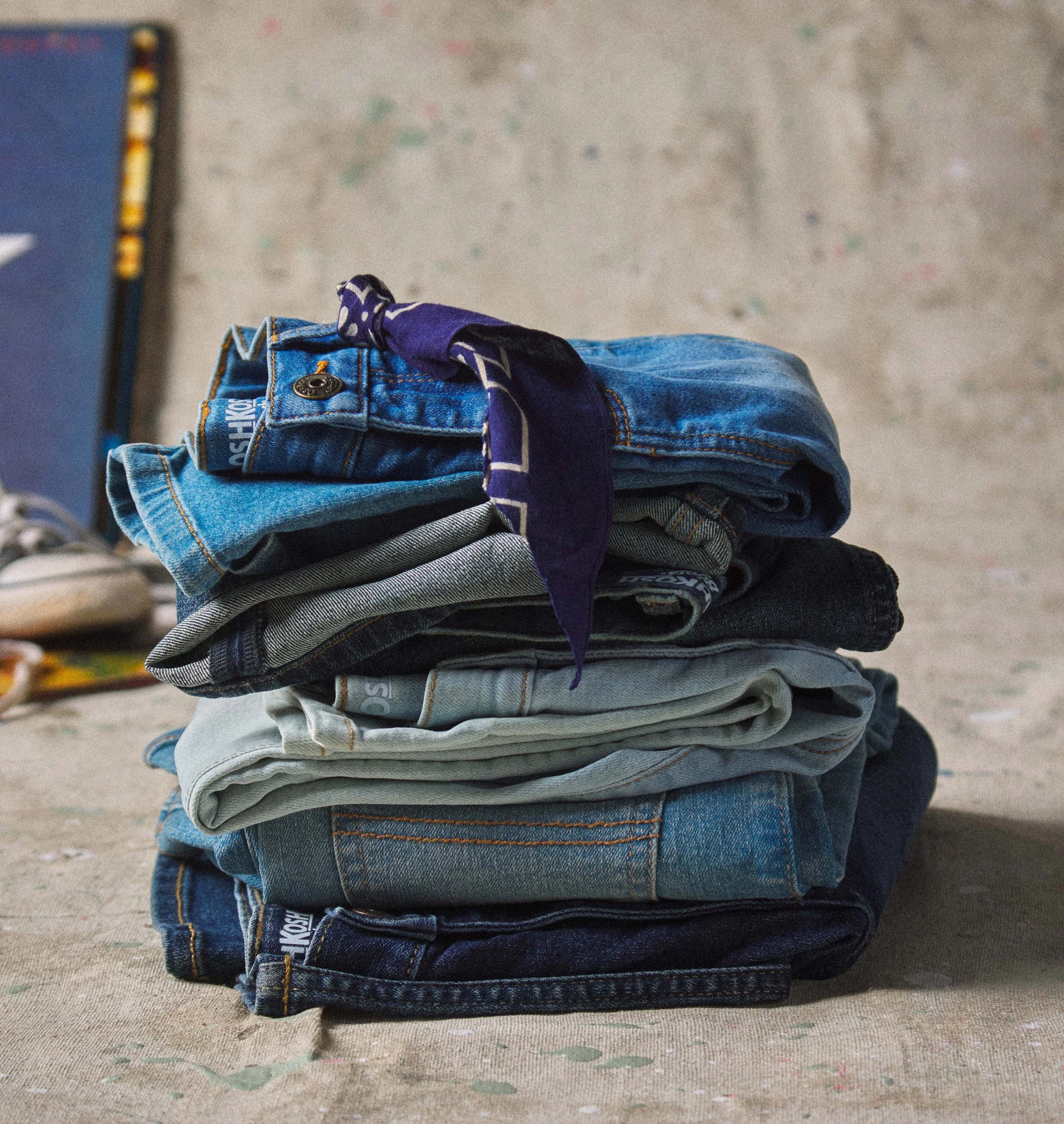 Stacked jeans with a blue bandana tied around one of the top pairs, placed on a textured surface.