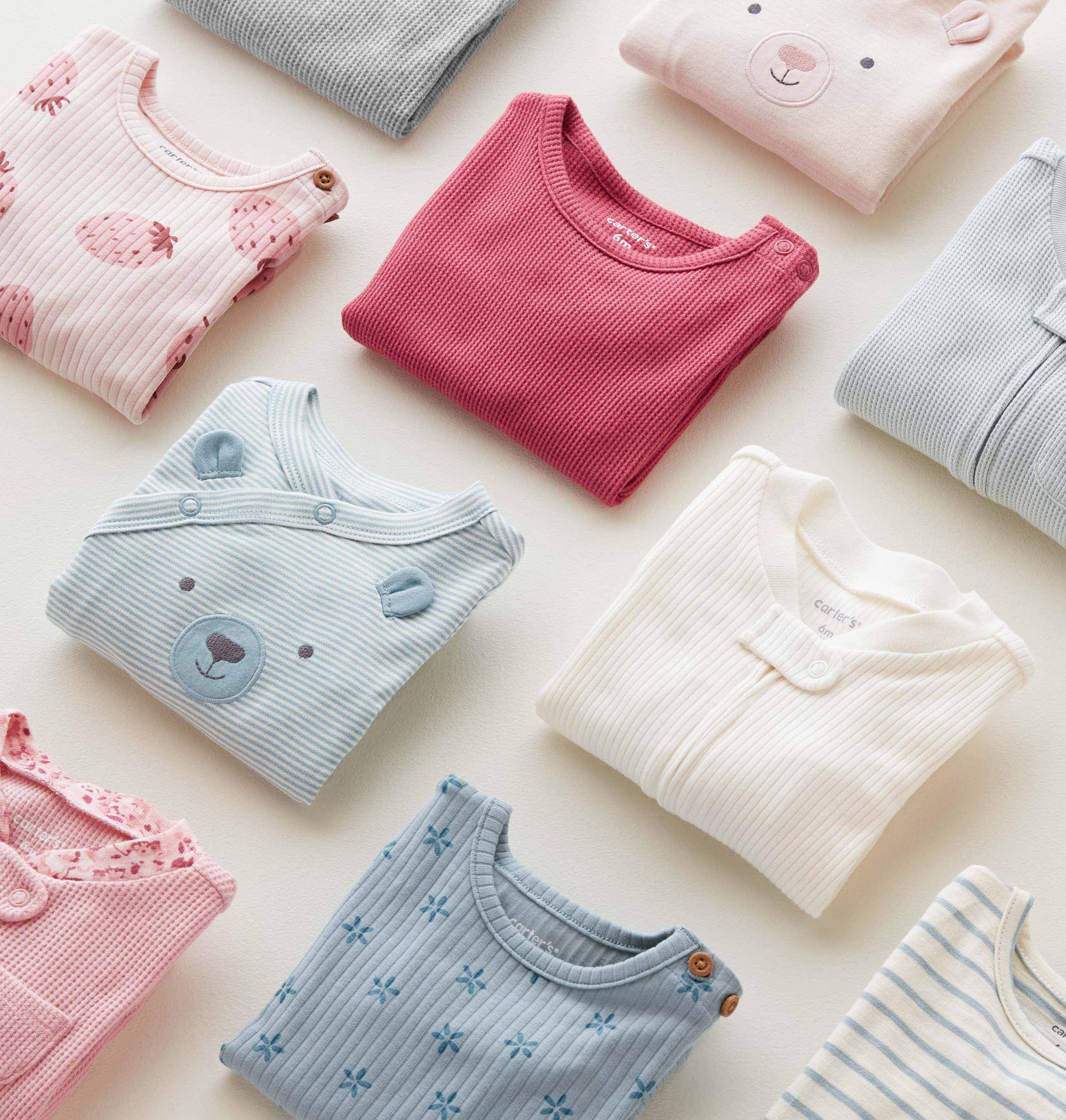 Baby clothes including a pink shirt with strawberry prints, a gray shirt, a pink shirt with bear face, a blue striped shirt, a white striped shirt, and a pink lace-trimmed outfit, arranged on a white surface.