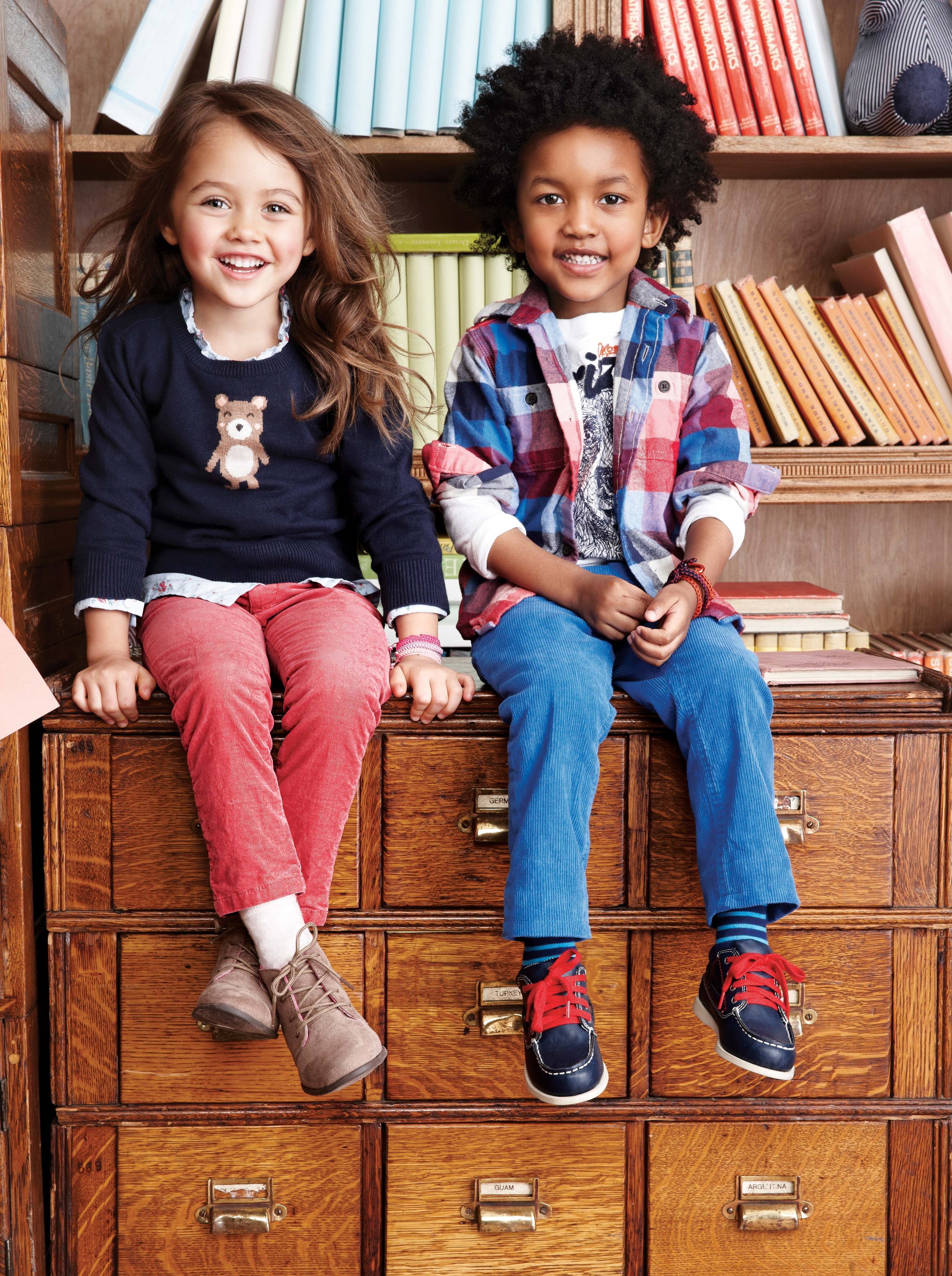Two children, a girl and a boy, sit on a wooden cabinet with labeled drawers. The girl has long brown hair, a navy sweater with a bear on it, red pants, and brown boots. The boy has curly black hair, a plaid shirt, blue pants, and blue sneakers with 