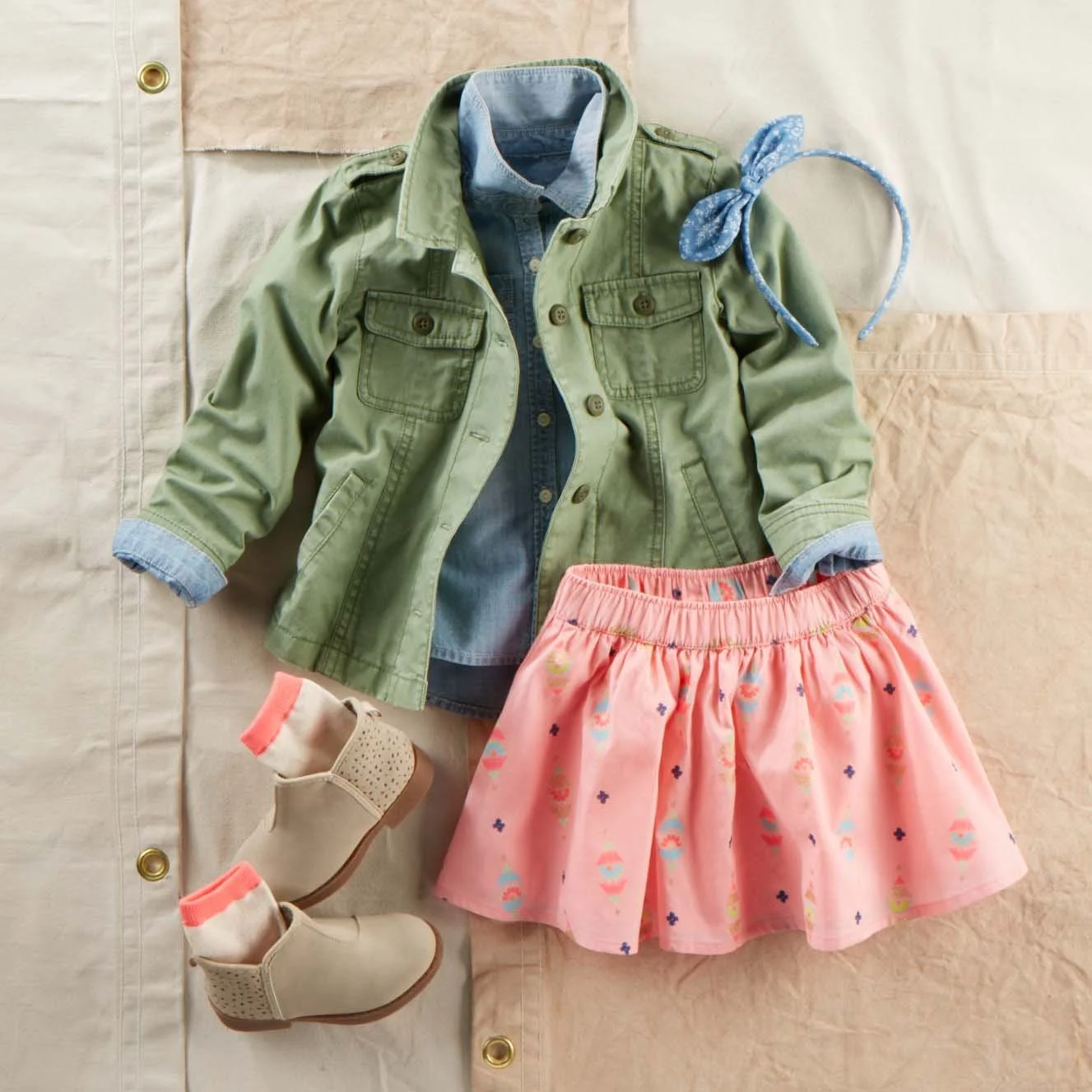 Children's clothing and footwear displayed on a surface, including beige ankle boots with pink accents, a pink skirt with colorful patterns, a denim shirt layered under an olive jacket, and a blue patterned headband.