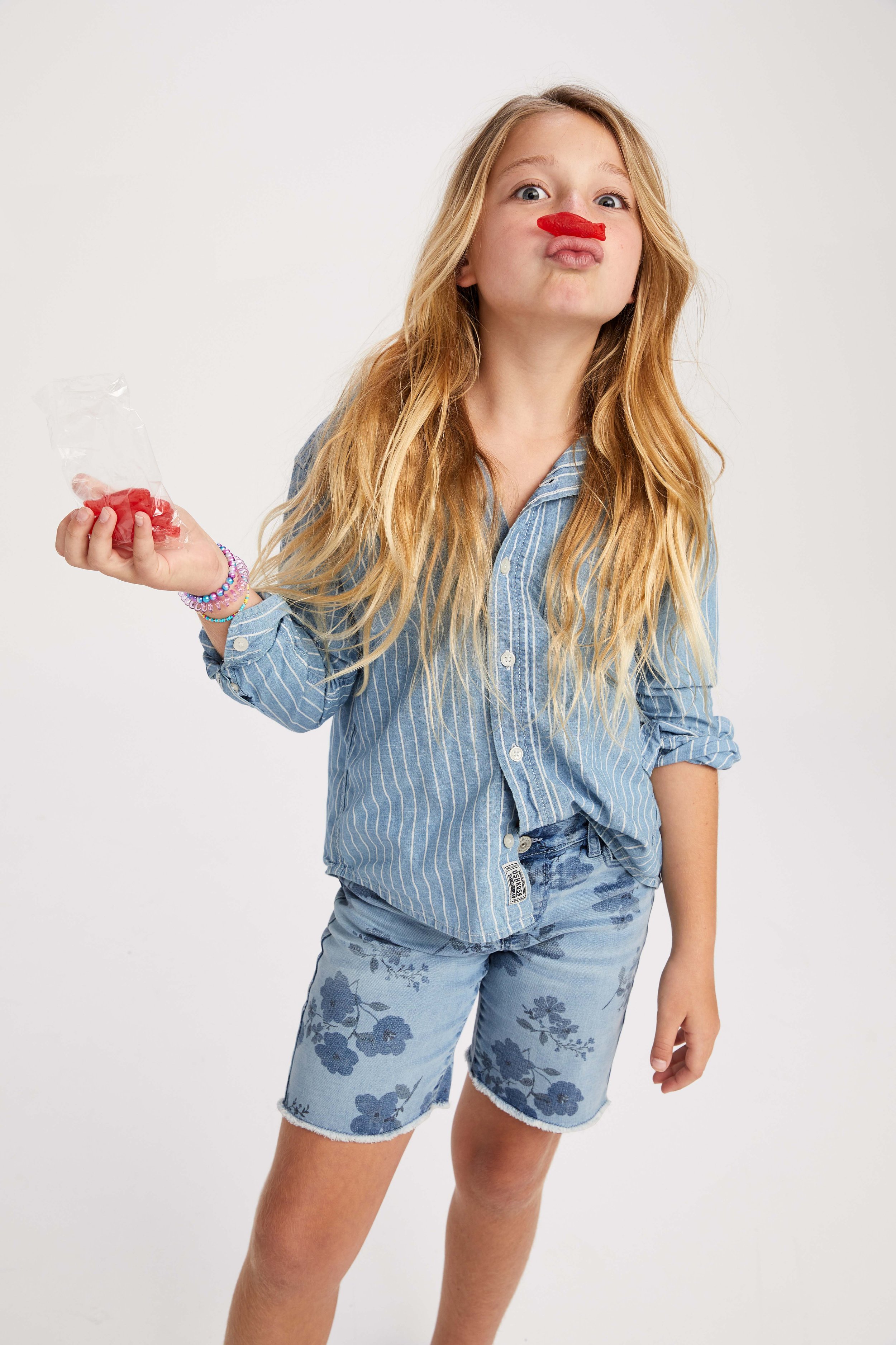 A young girl with long blonde hair and blue eyes making a funny face with her lips puckered and a red gummy candy on her nose, holding a bag of red gummy candies in her right hand, wearing a blue striped shirt and shorts with floral pattern against a