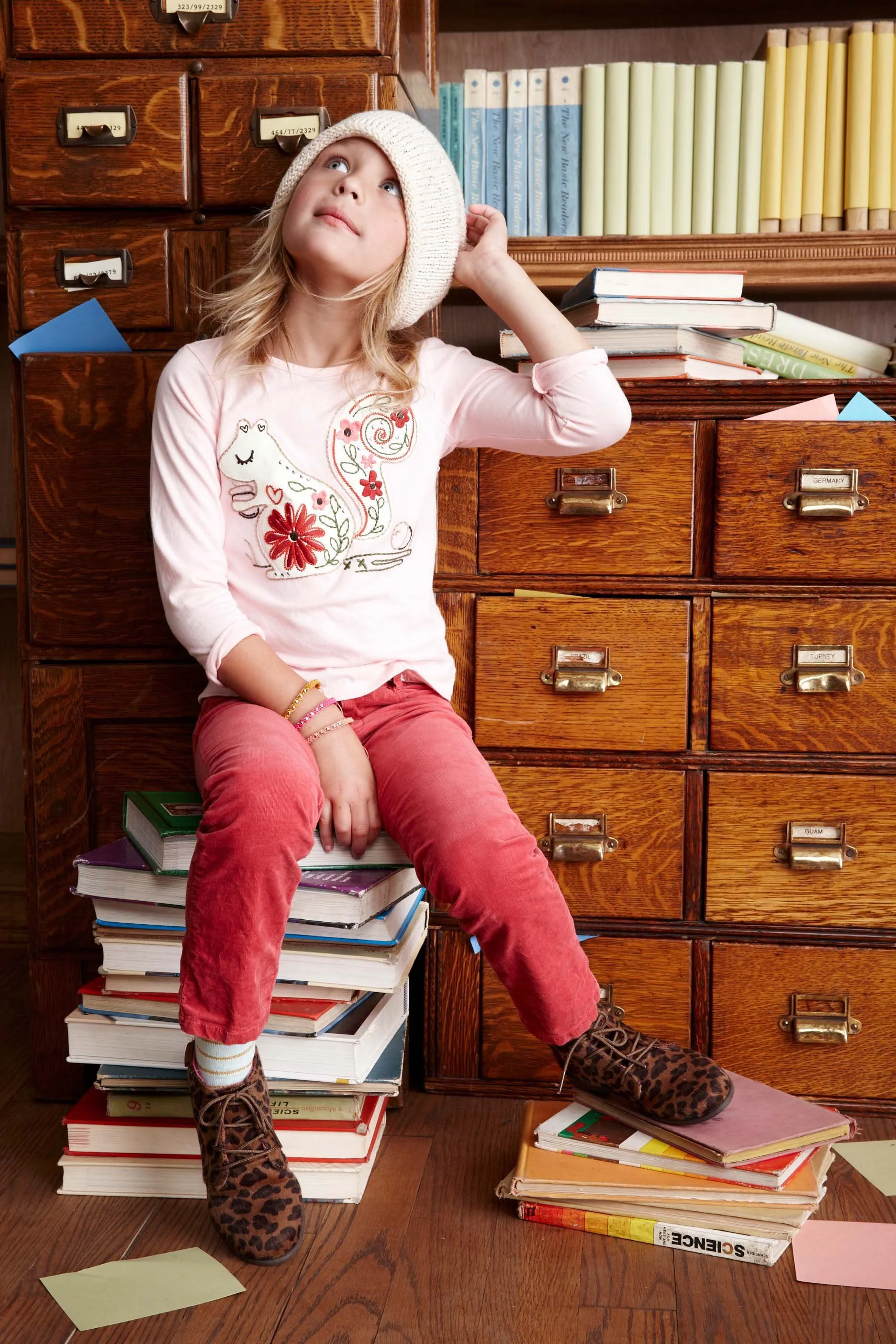 A young girl with blonde hair wearing a knit hat and a pink shirt, sitting on a stack of books in front of wooden drawers, surrounded by scattered papers and books, with bookshelves in the background.