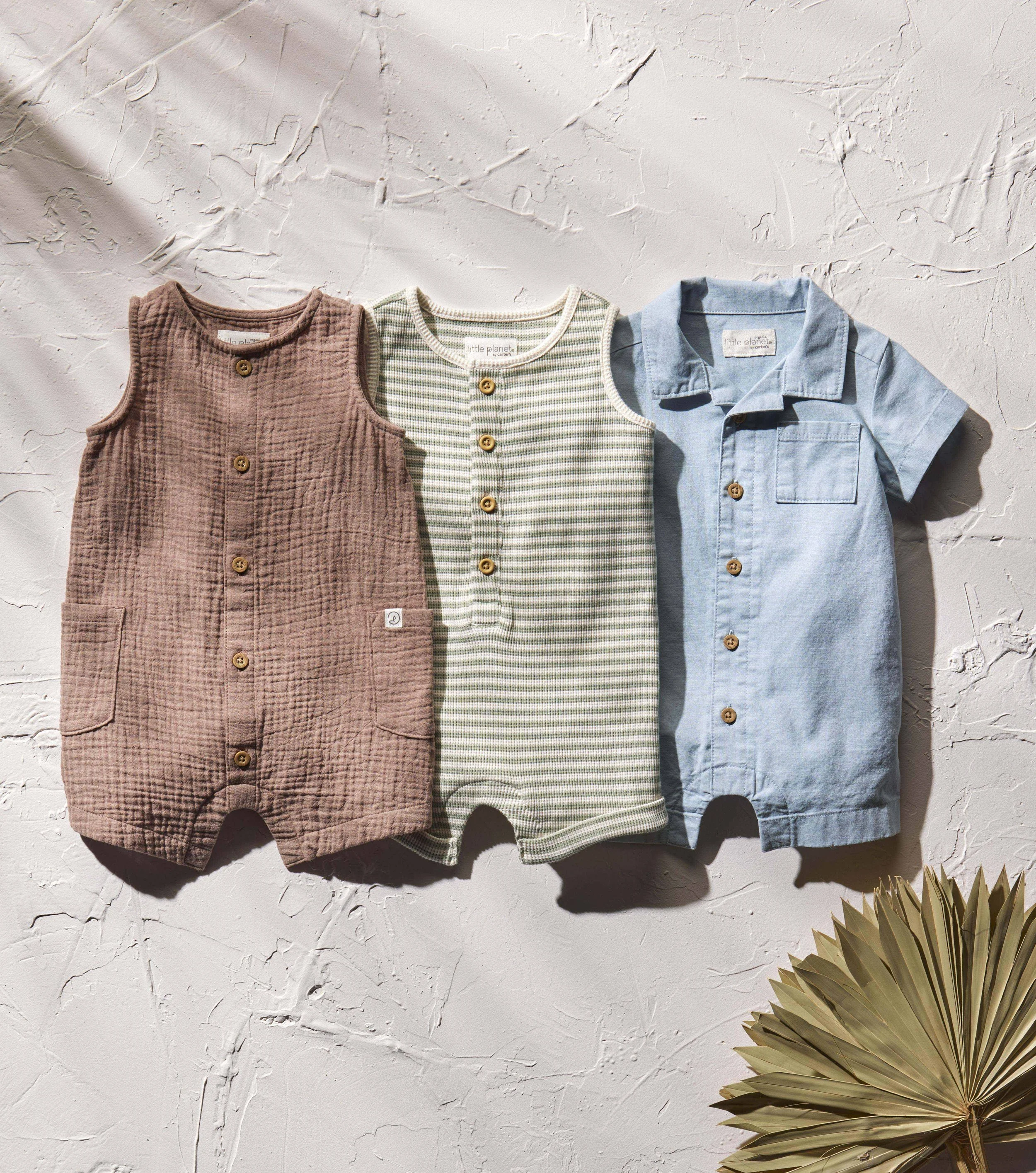 Three children's clothing items hang on a textured white wall with a dried palm plant in the bottom right corner. The clothes include a brown quilted romper, a sleeveless striped top, and a light blue shirt.