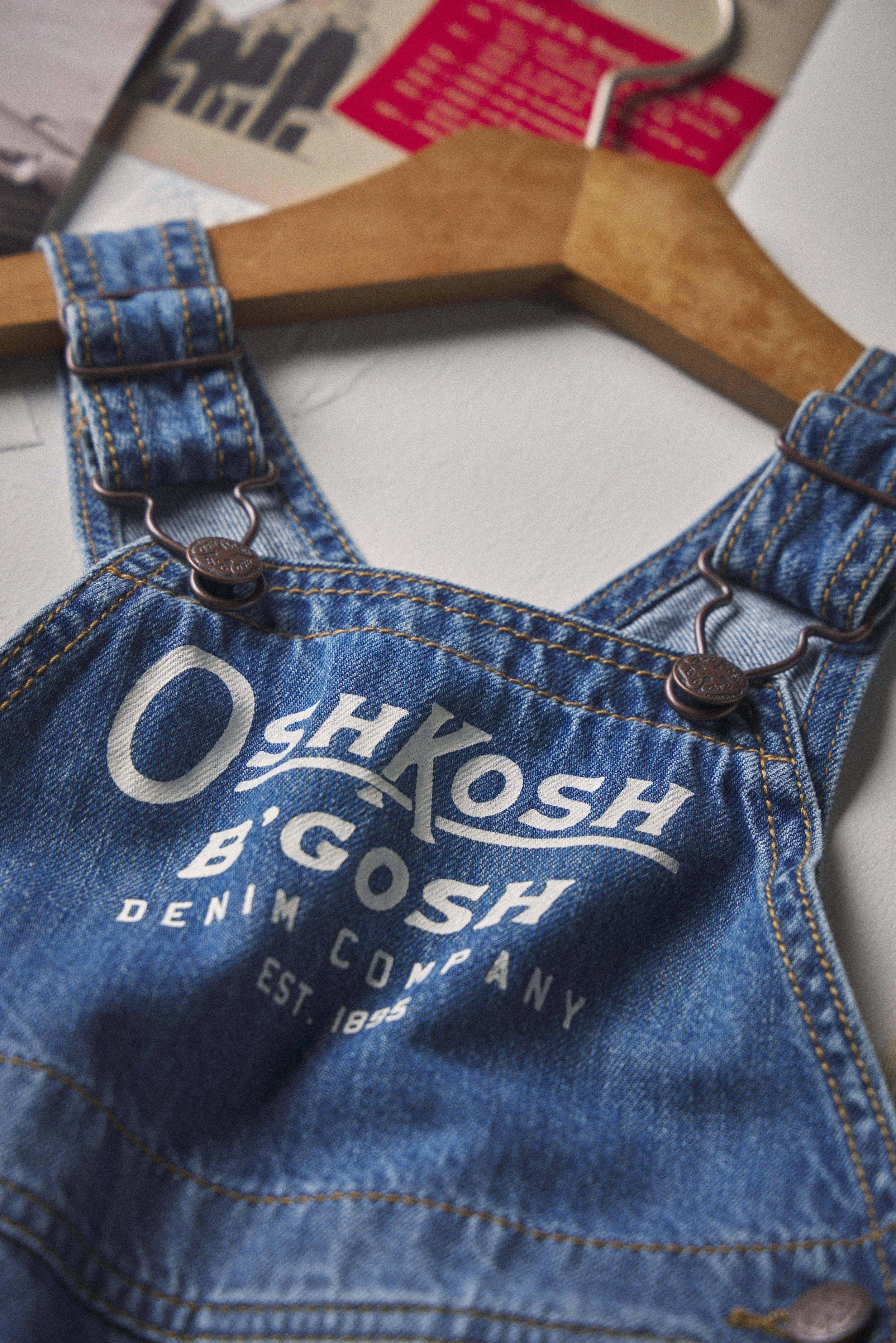 A close-up of a denim overall featuring white text and design, positioned on a hook hanger against a background of a magazine or newspaper.