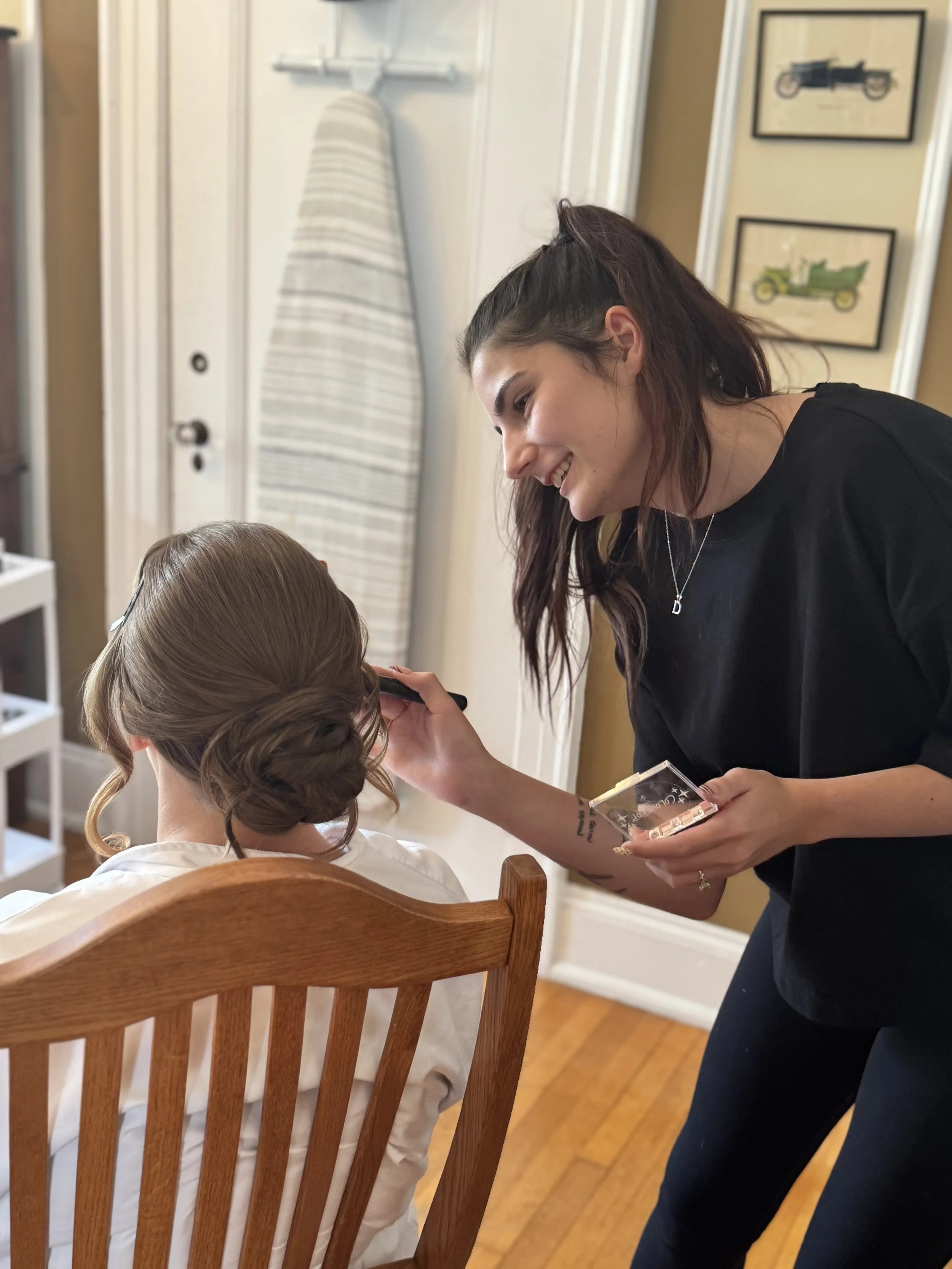 A woman with dark hair styled in loose waves, wearing a black t-shirt and necklace, is smiling and talking to a seated girl with brown hair styled in a braid, who is sitting in a wooden chair. The room has framed pictures of vintage cars on the wall and a closed door with a striped cloth cover hanging on a hook.