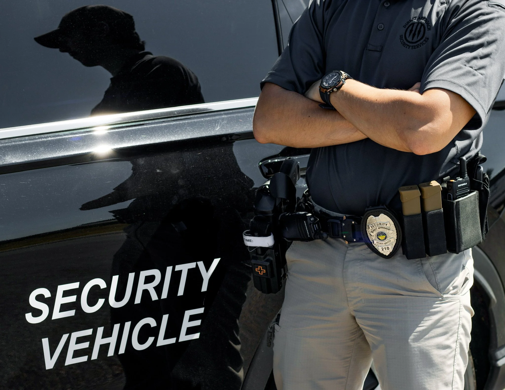 A security officer with crossed arms standing beside a black vehicle with a security agency logo. The officer is wearing a gray uniform with a security badge and a wristwatch. The vehicle has the words 'Security Vehicle' visible on it.