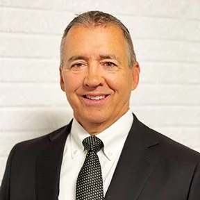 Portrait of a smiling man in a black suit, white shirt, and dotted tie, standing in front of a white brick wall.