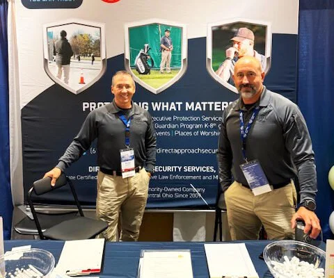 Two men standing behind a table at a conference booth, smiling. The booth has a backdrop with images of outdoor scenes and text about protective services.