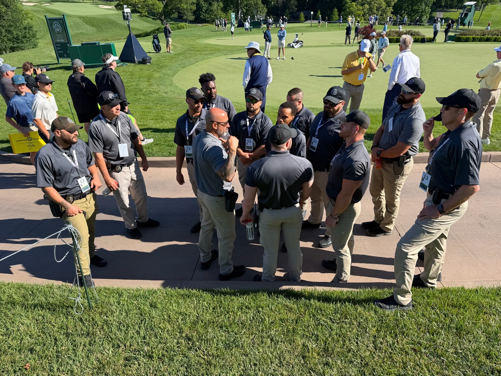 Group of people gathered near the putting green at a golf tournament, with some players, officials, and spectators, on a sunny day at a golf course.