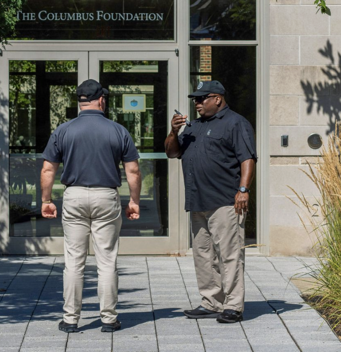 Two security guards talking outside a building with the sign 'The Columbus Foundation' on the door. One guard is facing away from the camera, and the other is facing slightly to the right, holding a walkie-talkie.