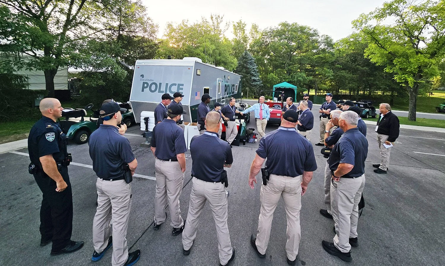 Group of police officers and officials gathered outdoors in a circle, with police vehicles and golf carts nearby, during a meeting or briefing in a parking lot surrounded by trees in the evening.
