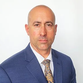 A professional man wearing a blue suit, white shirt, and patterned tie, posing against a plain white background.