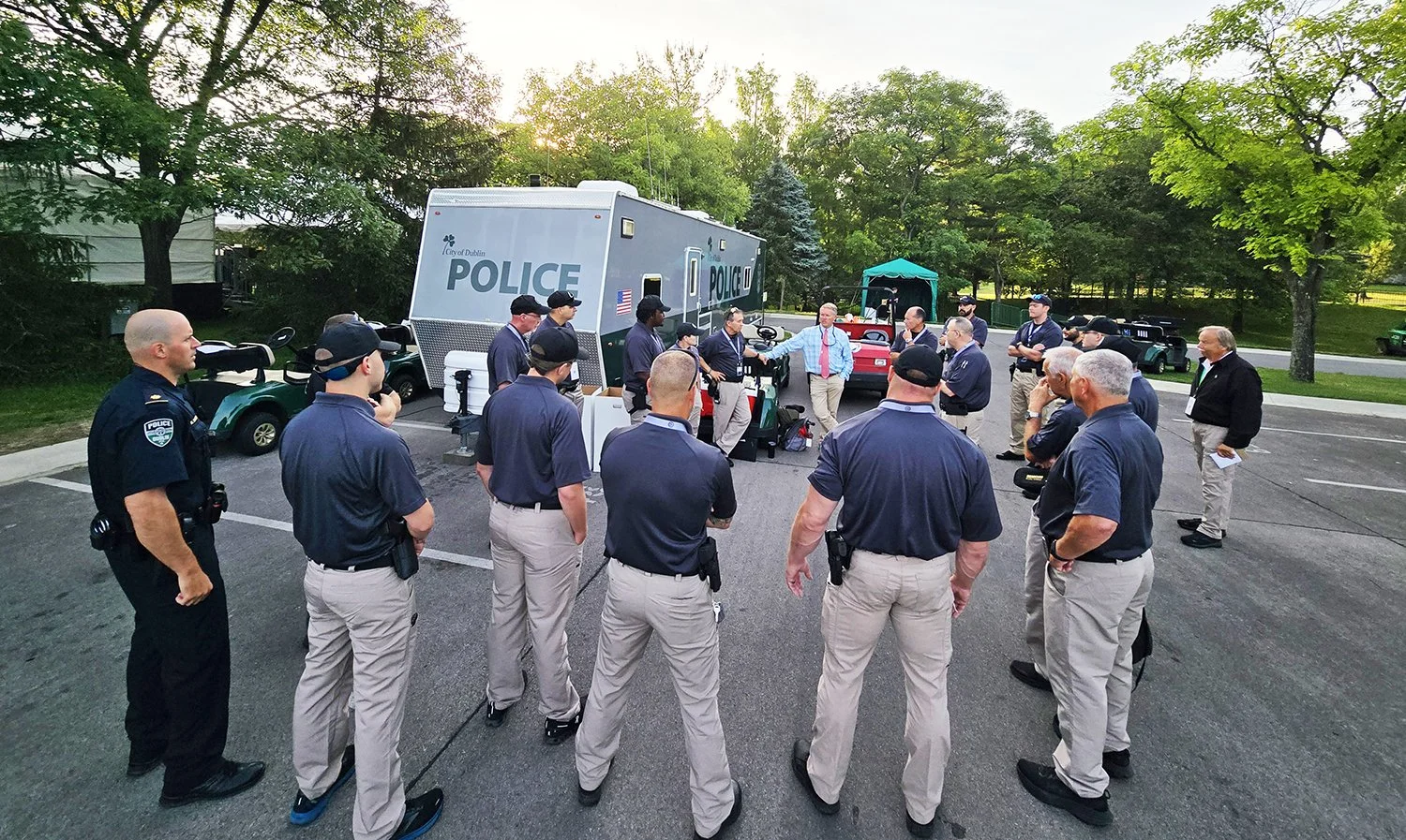 Group of police officers and other individuals standing around in a parking lot, near a police mobile command center, with golf carts and trees in the background, during sunset.