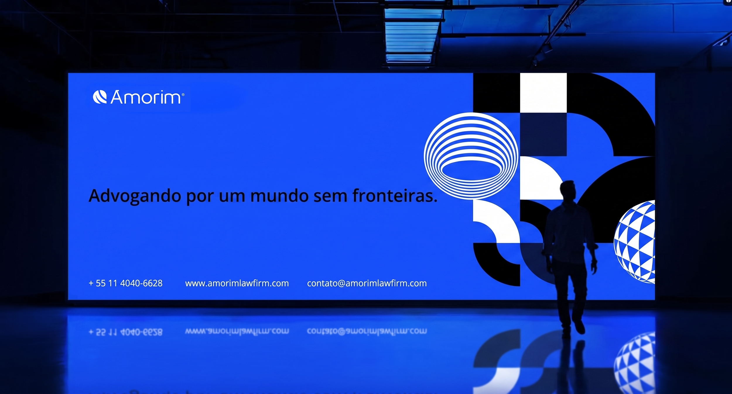Painel com fundo azul e elementos gráficos geométricos em cores preto, branco e azul. Texto em português 'Advogando por um mundo sem fronteiras'. Logo 'Amorim' no canto superior esquerdo, informações de contato na parte inferior.