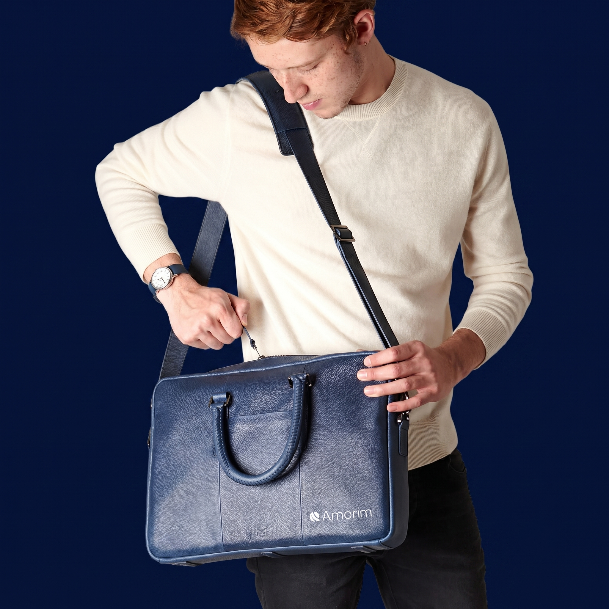 Pessoa com cabelo castanho claro, vestindo suéter branco, usando relógio e carregando uma bolsa azul escura com alças pretas, contra fundo azul escuro.