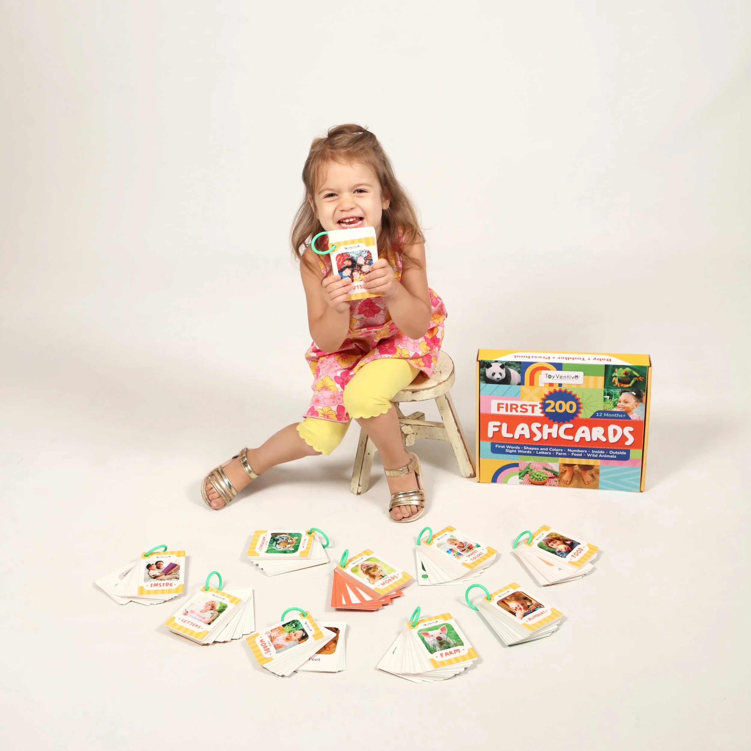 Menina sorridente sentada em um banquinho segurando um cartão de flashcards colorido, com uma caixa de flashcards ao lado e outros cartões espalhados no chão.