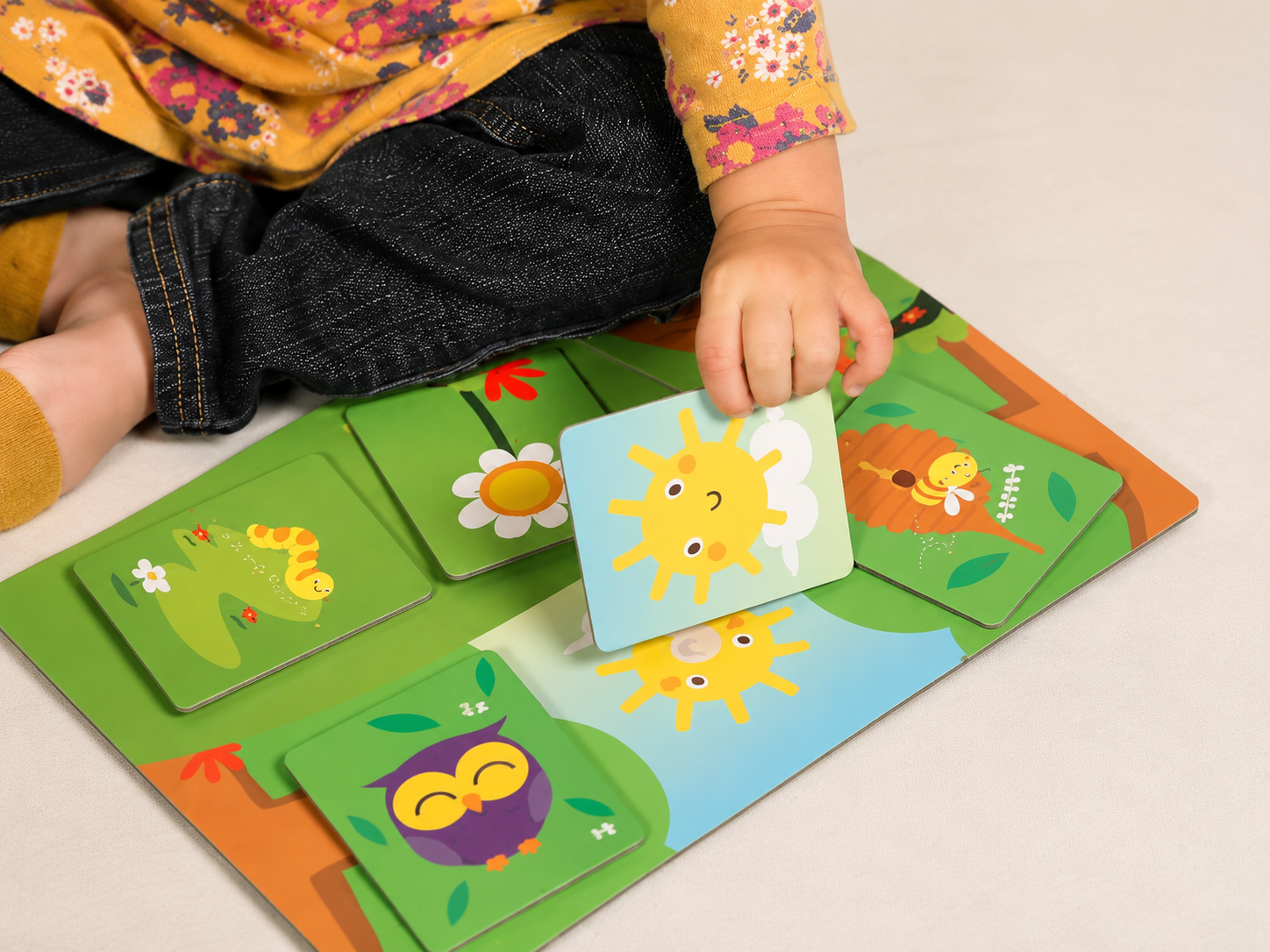 Criança segurando uma carta de um jogo de faz de conta com tema de animais, no tabuleiro há imagens de uma lagarta, uma girafa, uma abelha, uma coruja e uma flor, tudo com cores vibrantes e um tema natureza.
