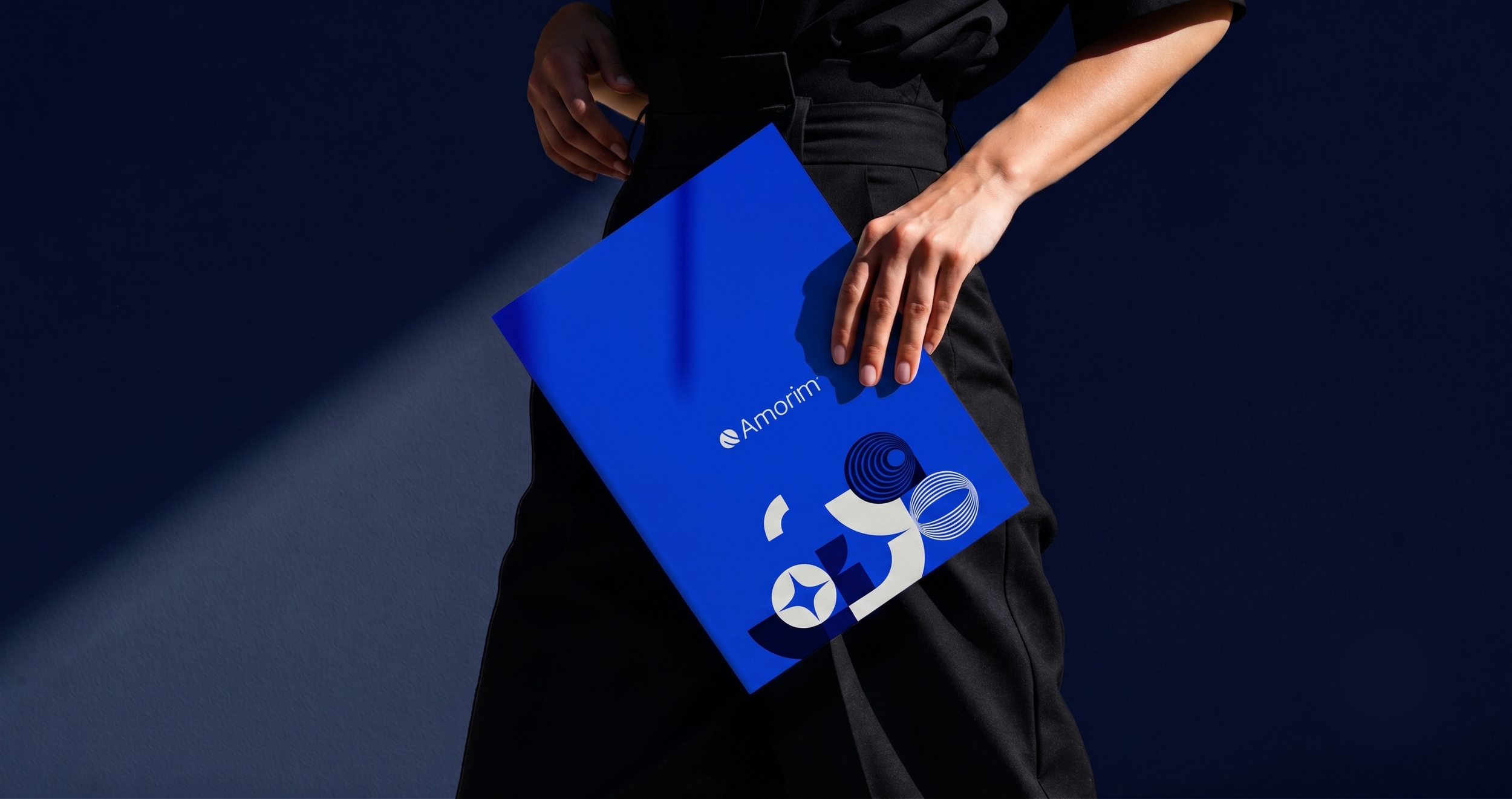 Pessoa segurando uma pasta azul com o logotipo da Amorim, vestindo roupas pretas, contra um fundo escuro com iluminação ressaltando a pasta.