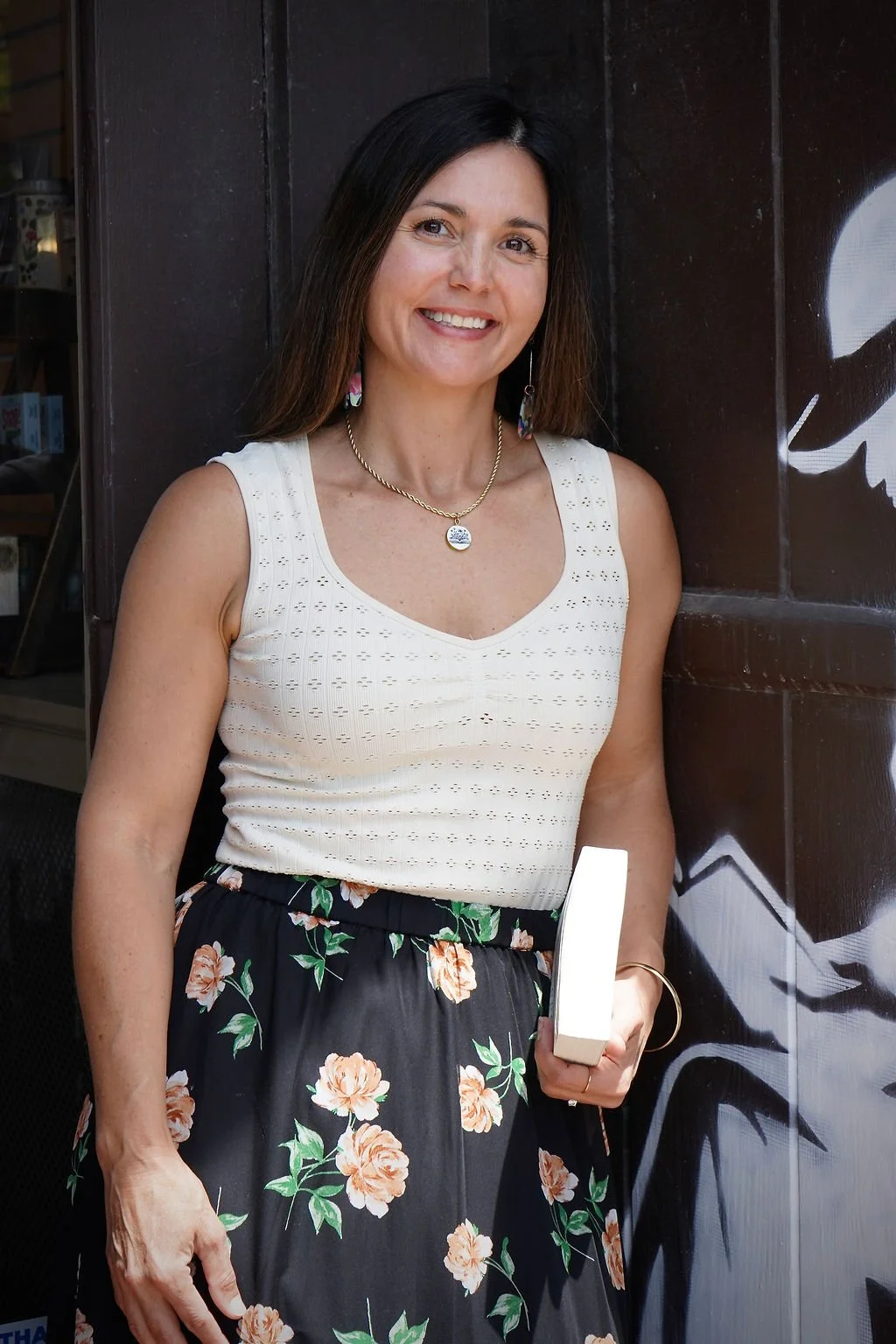 A woman with dark brown hair smiling, wearing a white sleeveless top, a black floral skirt, and jewelry, standing next to a black wooden wall with graffiti.
