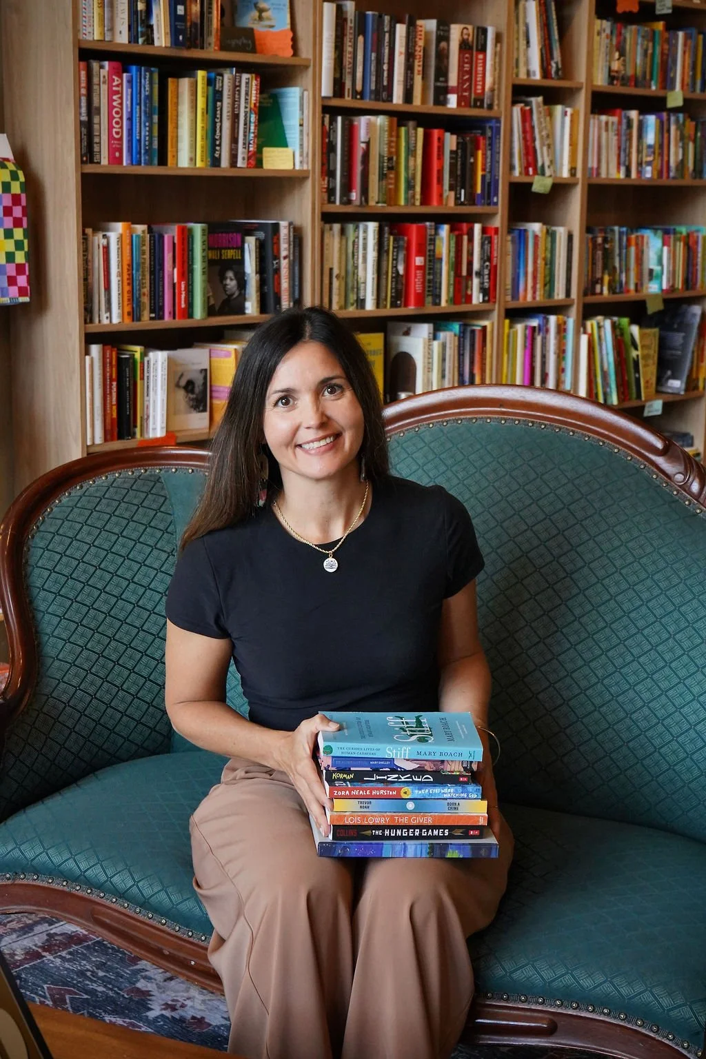 A woman with long dark hair sitting on a vintage green upholstered sofa in a library, holding a stack of books, with bookshelves filled with colorful books in the background.