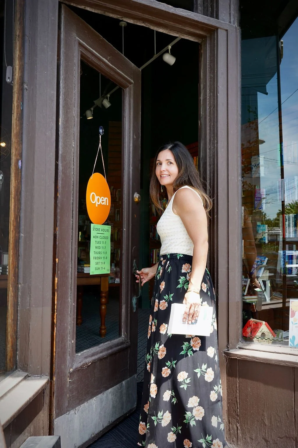 A woman with long dark hair, wearing a white sleeveless top and a long floral skirt, is opening the door of a bookstore or library. She is holding a white book or notebook in her left hand and smiling at the camera. The store has an "Open" sign hanging on the door and store hours listed on a green paper.