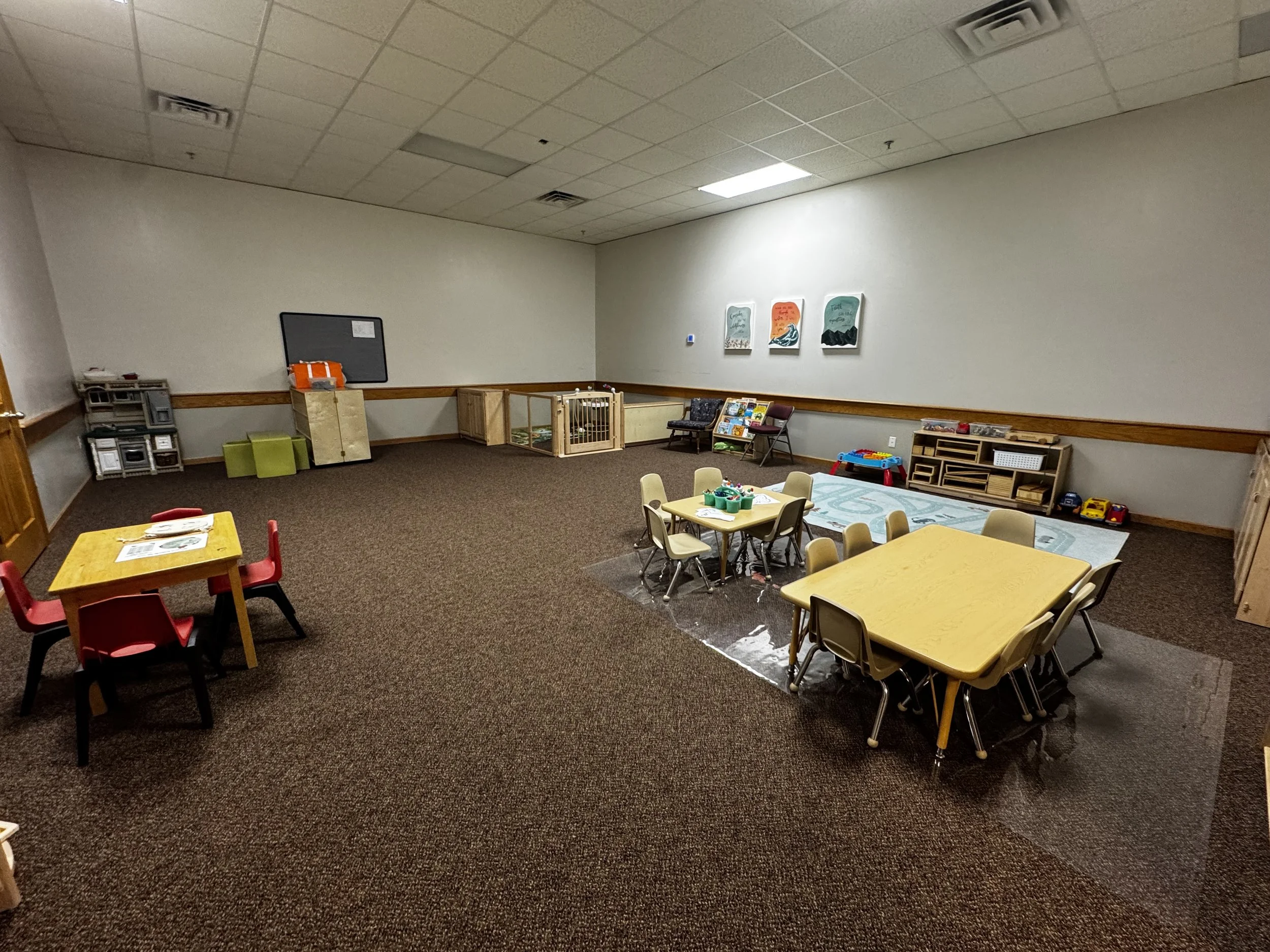 A room with small tables and chairs, children's toys, books, and wall art, indicating it is a classroom or playroom for young children.
