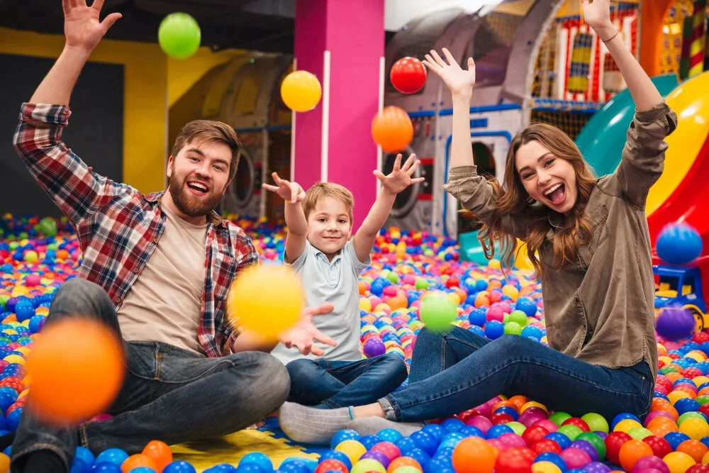A smiling family of three playing in a colorful indoor ball pit, throwing balls in the air and laughing.