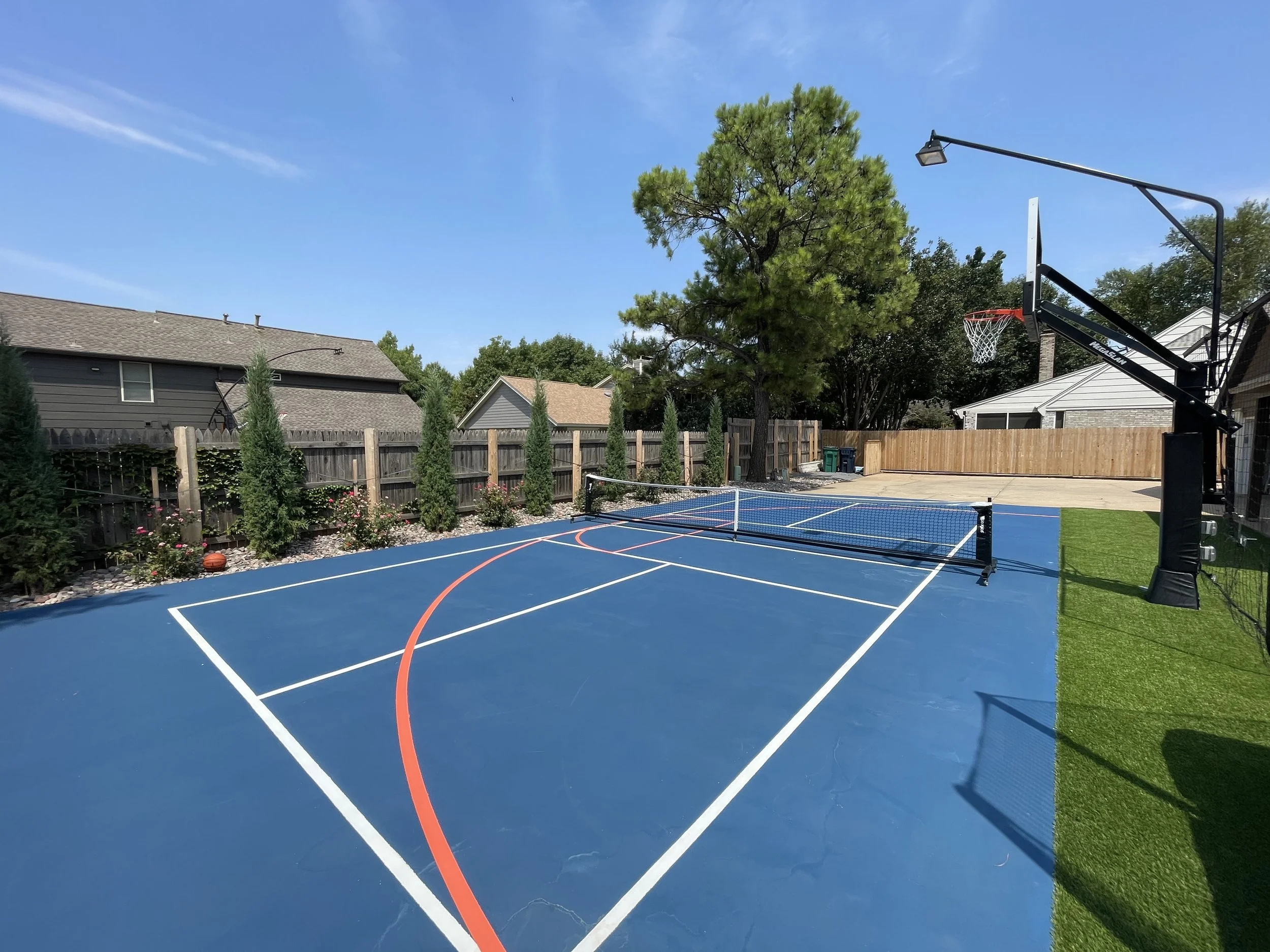 A backyard with a basketball court with blue surface, white and red lines, a basketball hoop, green grass, trees, and houses under a clear blue sky.