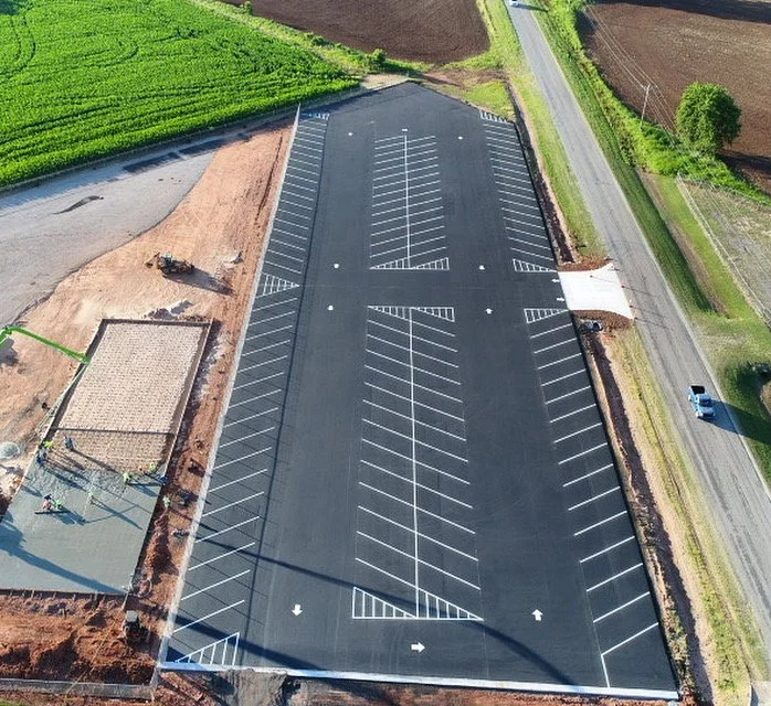 Empty parking lot under construction with designated parking spaces, surrounded by fields and a road.