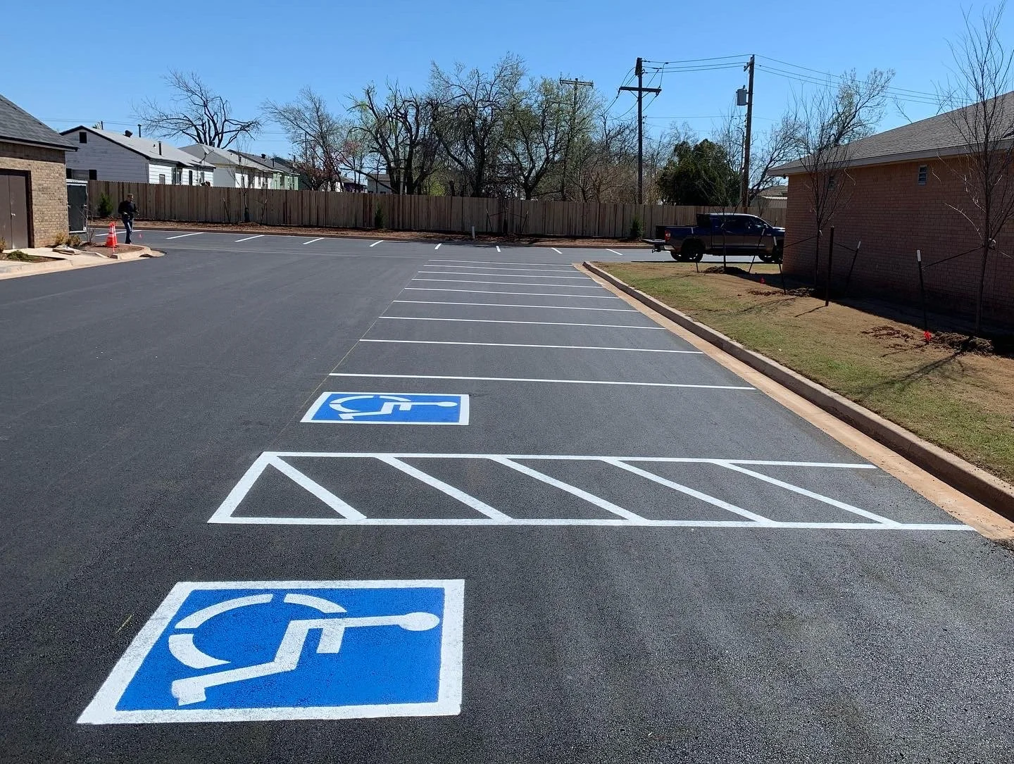 Empty parking lot with two accessible parking spaces marked in blue and white, close to a brick building, and a black pickup truck parked further away. Clear sky and leafless trees in the background.