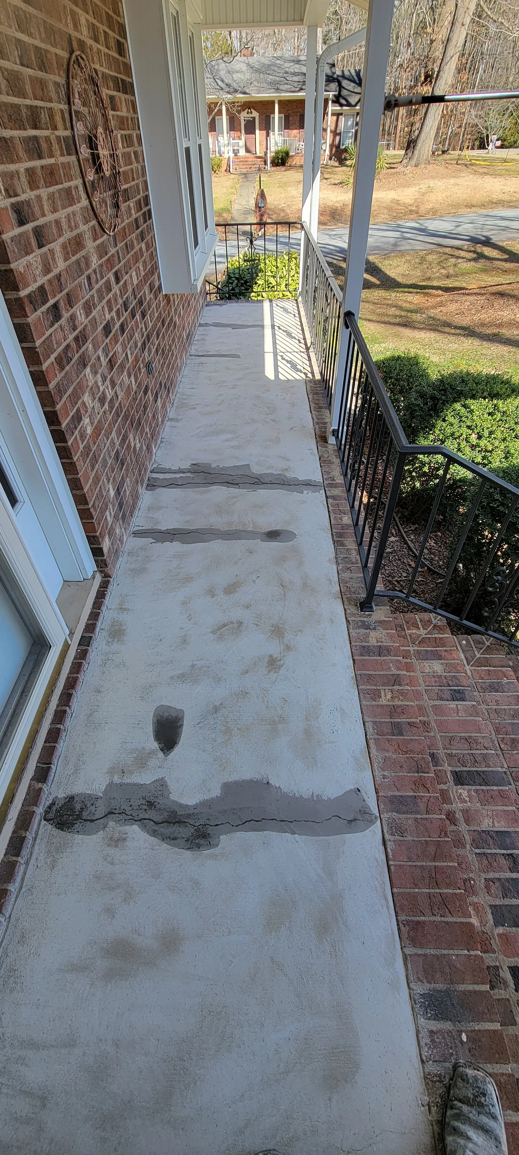 View of a porch with a concrete walkway and brick accents, showing wet footprints on the concrete and a brick house in the background.