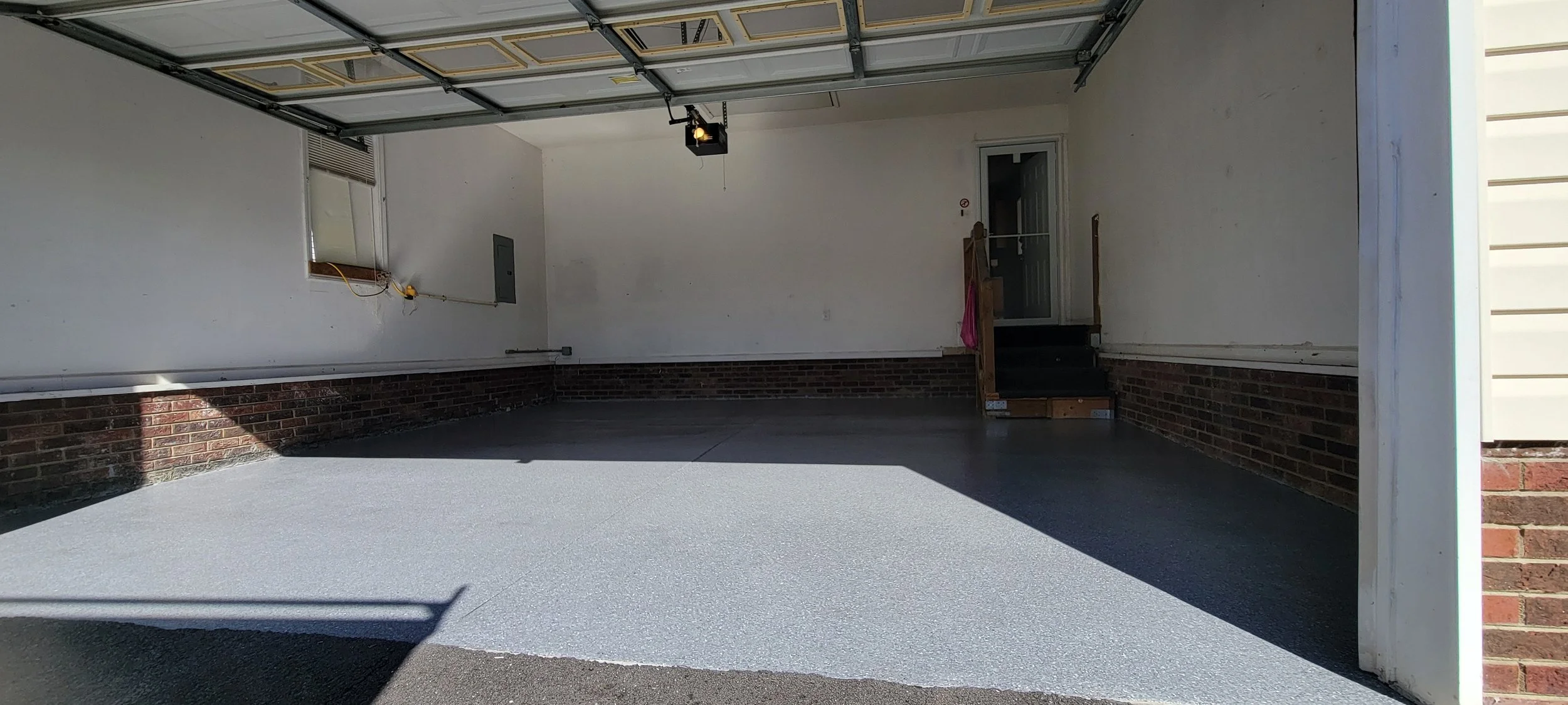 Empty garage with a textured gray floor, white walls, a brick wainscot, and a partially open automatic garage door.