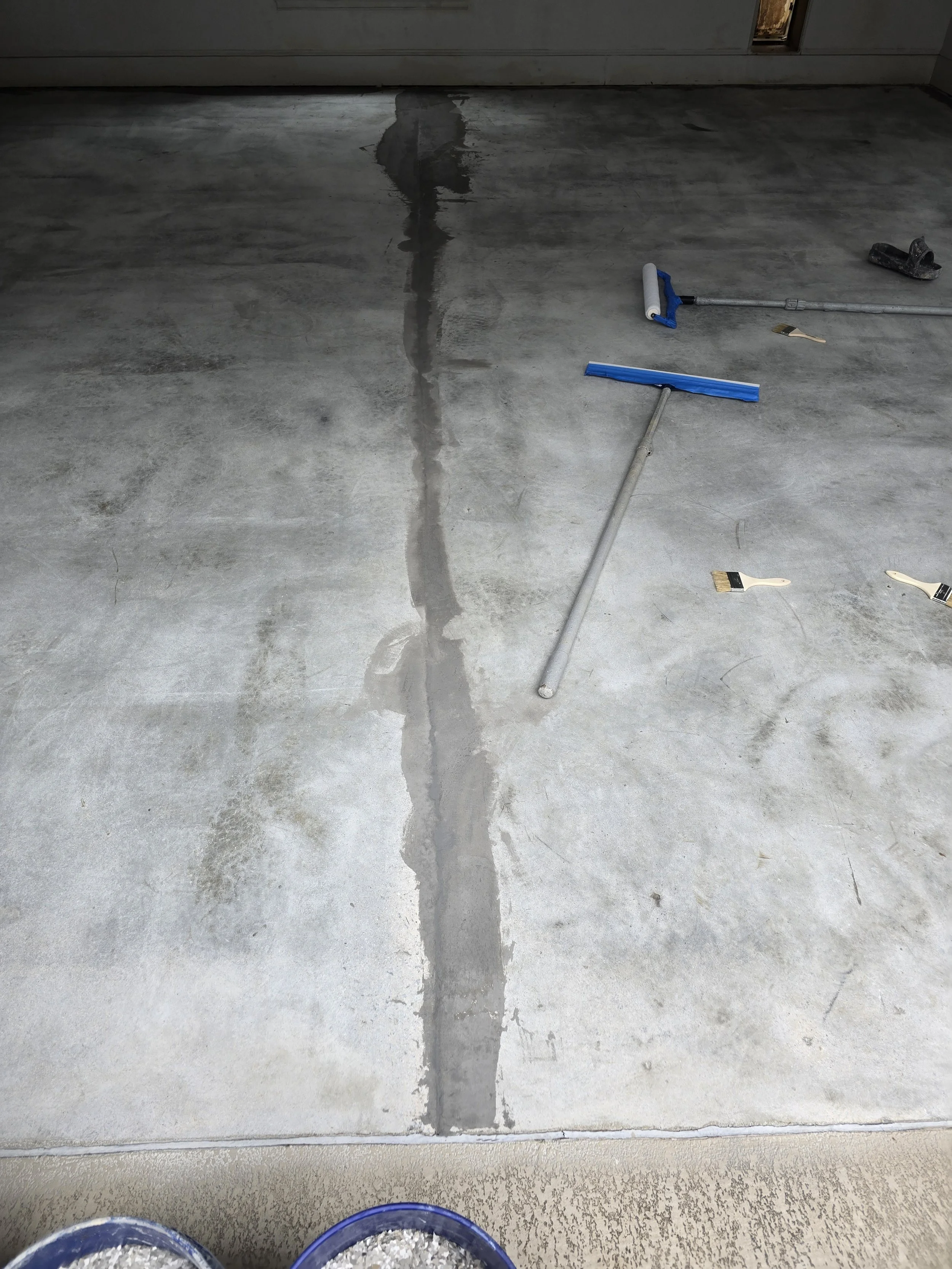 Concrete floor with a crack, construction tools including a floor scraper, brushes, and a blue container.