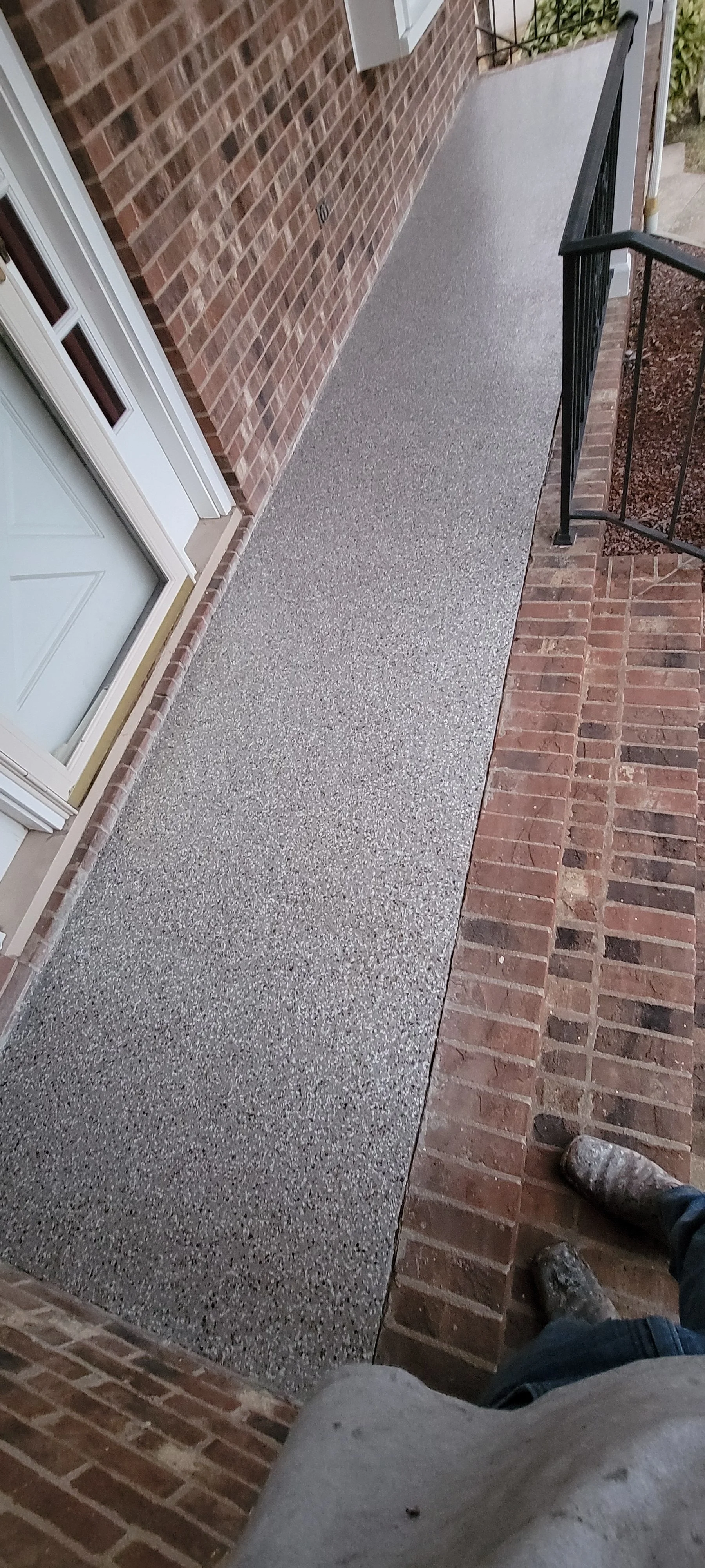 A front porch with a textured concrete walkway, a black metal railing, a brick wall, and a white door with glass panels.