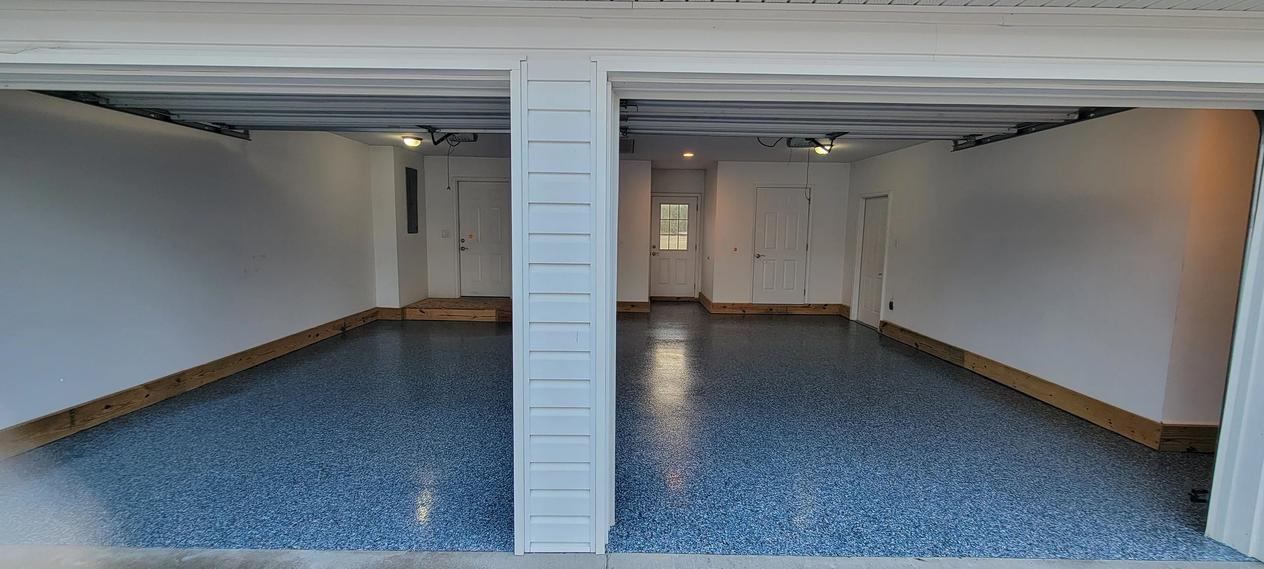 Two-car garage with white walls, wooden trim, gray speckled flooring, and ceiling lights, with a garage door open.