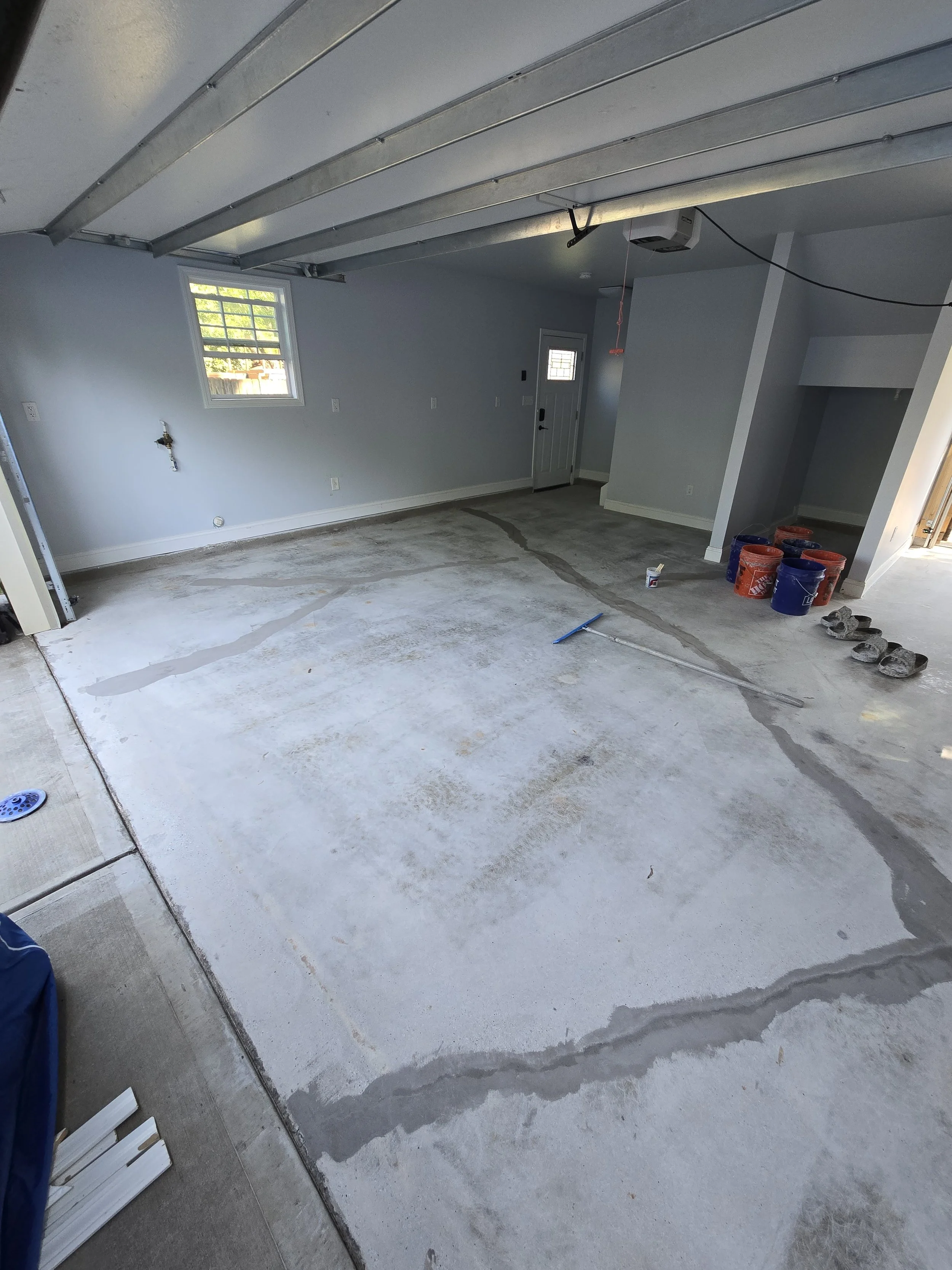 A garage interior under construction with an unfinished concrete floor, a window with a view of trees outside, and various construction materials including buckets and slippers.