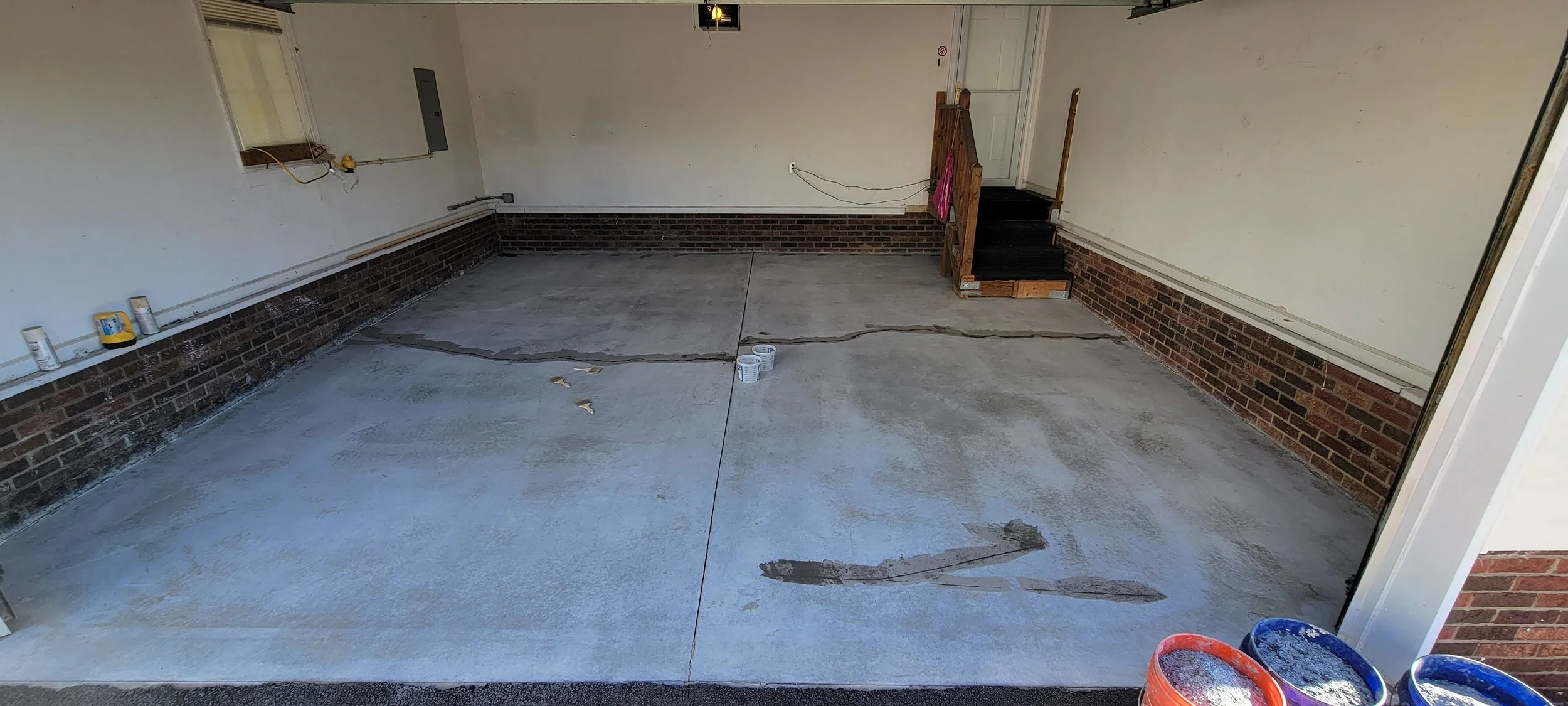A garage with a concrete floor being prepared for painting or sealing, leaning against the wall are cans of paint, and on the wall are bricks and pipes.