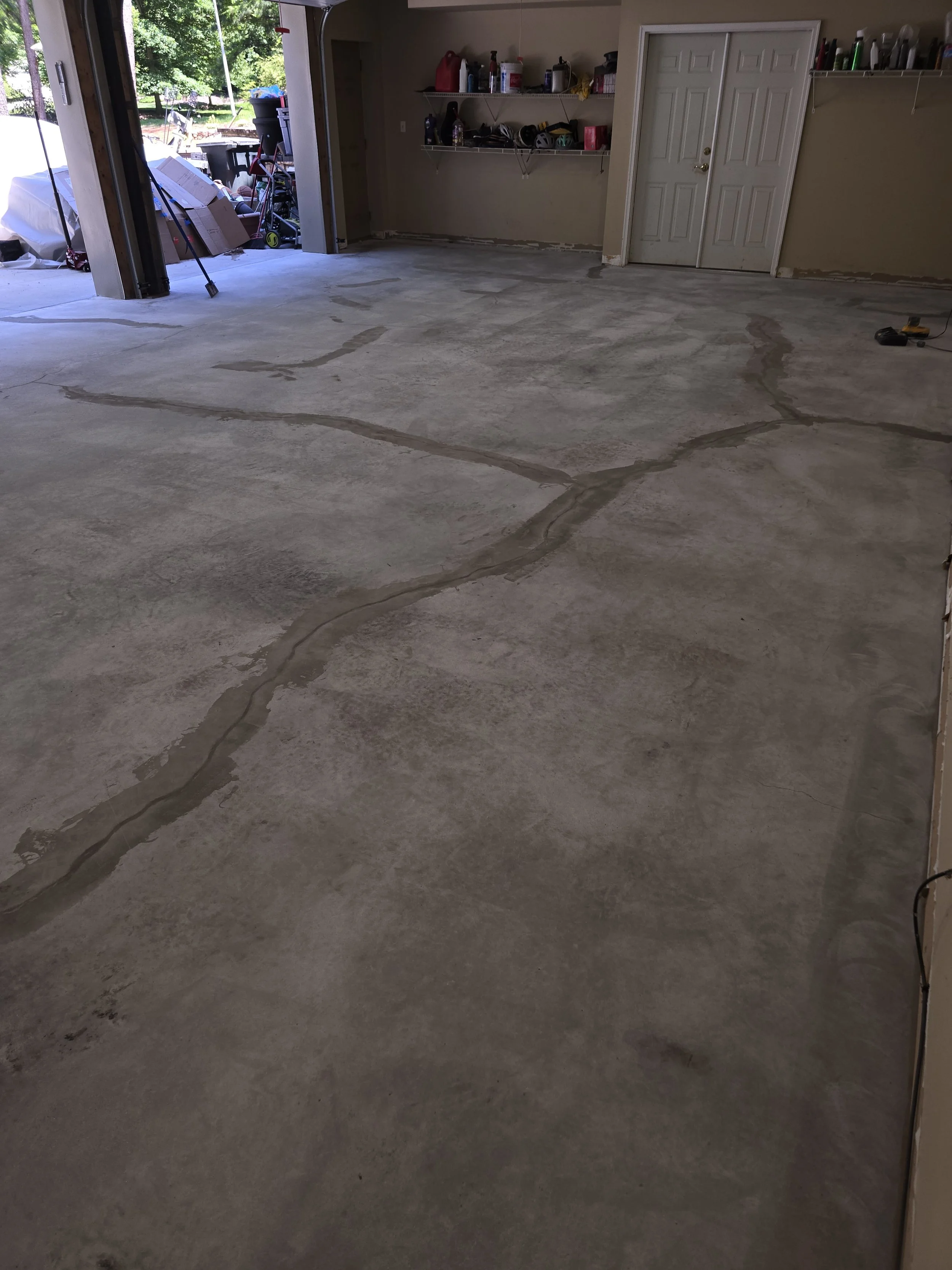 An empty garage with a concrete floor that has visible cracks and wet patches. The garage door is open, revealing a yard with trees, chairs, and outdoor items. Inside, there is a closed white double door, shelves with various items, and some tools on the floor.