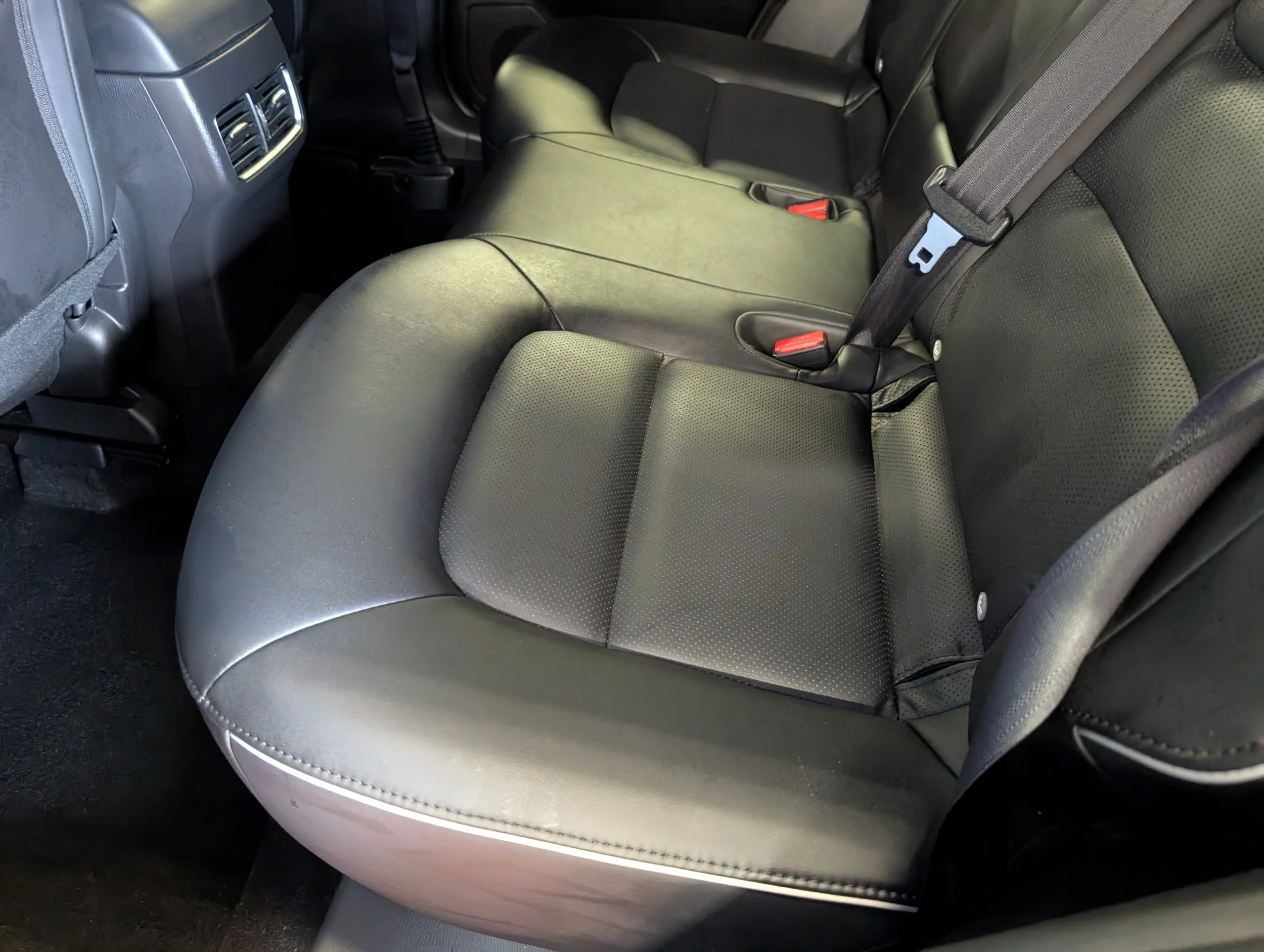 The backseat interior of a car, featuring black leather seats with visible seat belts, air vents, and part of the vehicle's rear side panel.