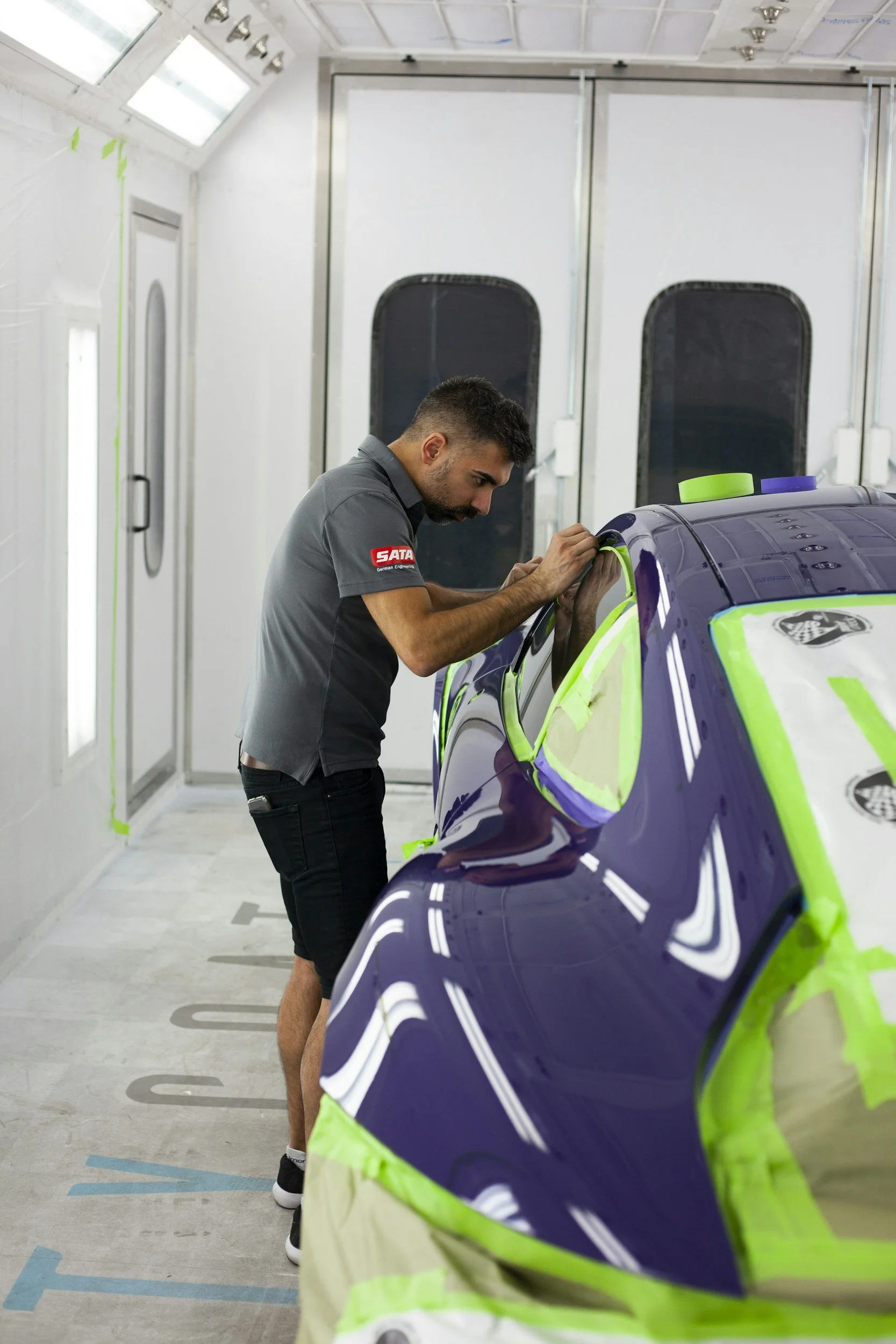 A man working on a purple and green race car inside a white spray booth, wearing a gray shirt with a red patch, painting the car.