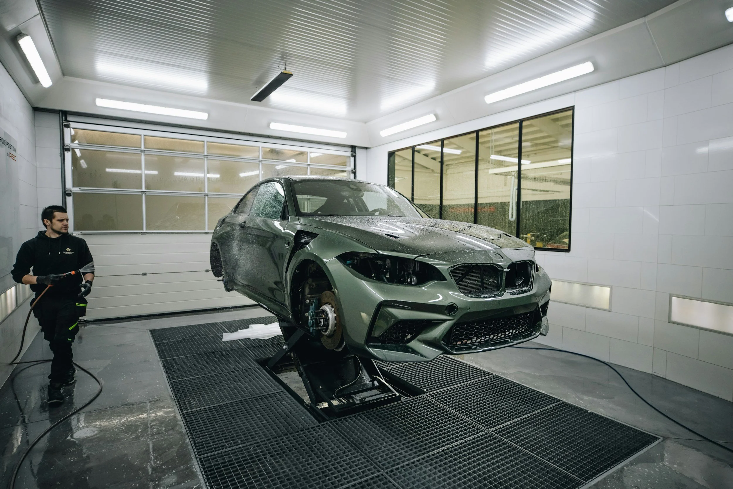 A person washing a silver sports car inside a well-lit auto detailing shop with white walls, large windows, and a grated floor.